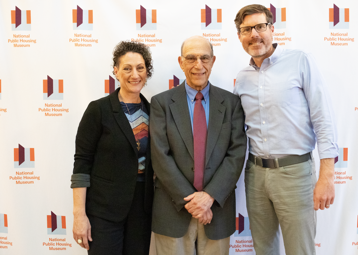  Authors Richard and Leah Rothstein stand next to co-founder of btcRE, M. Ryan Gorman, the sponsor of the event. They all stand in front of a branded white banner with small National Public Housing Museum logos repeated across it.  