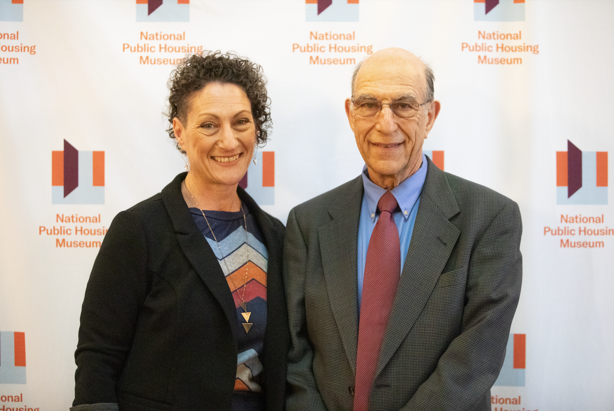  Authors Richard and Leah Rothstein stand in front of a branded white banner with small National Public Housing Museum logos repeated across it.  