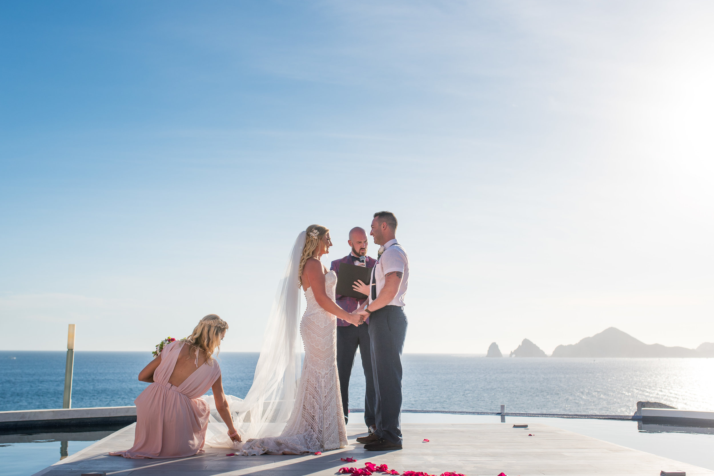 Wedding ceremony at Sunset Monalisa. Bride and groom are holding hands and one of the bridemaids is fixing bride´s dress