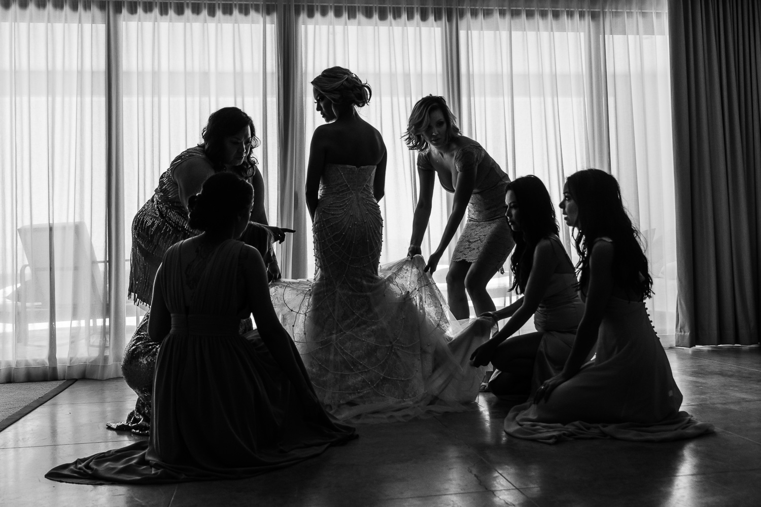  The picture beautifully captures from back the bride getting ready for her wedding day with all her bridesmaids. She is wearing an elegant white gown with intricate details on it and is getting it set with the help of a few of her bridesmaid- destin