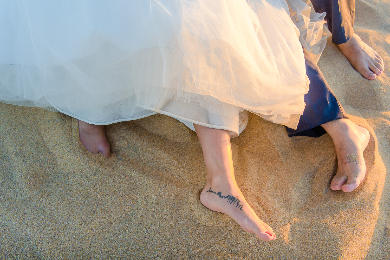  Bride and groom trash teh dress, both of them are layng down on the sand and barefoot. Bride has an interesting tatoo on her leg   