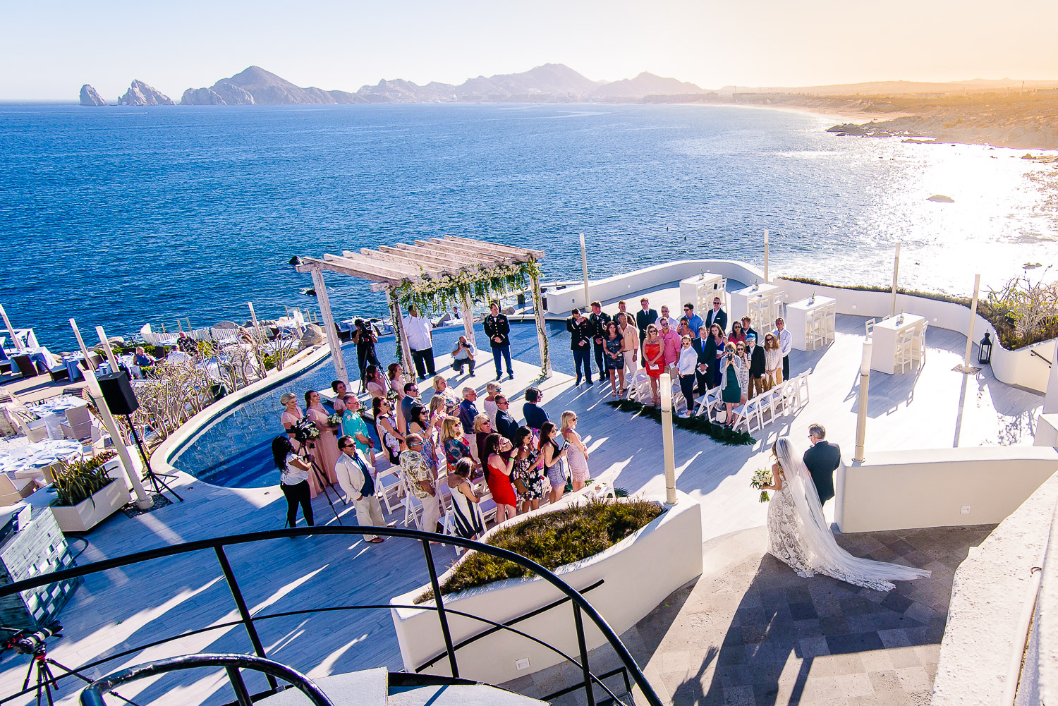  Aerial photo during a Los Cabos  wedding ceremony. Bride and father of the bride walking to the aisle. Blue ocean and famouse cabo arch as a background 