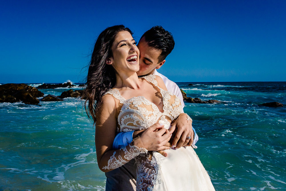 Bride and groom during her trash the dress session in Los Cabos. Both of them are laughing as the groom is holding his beautiful bride from behind on the beach