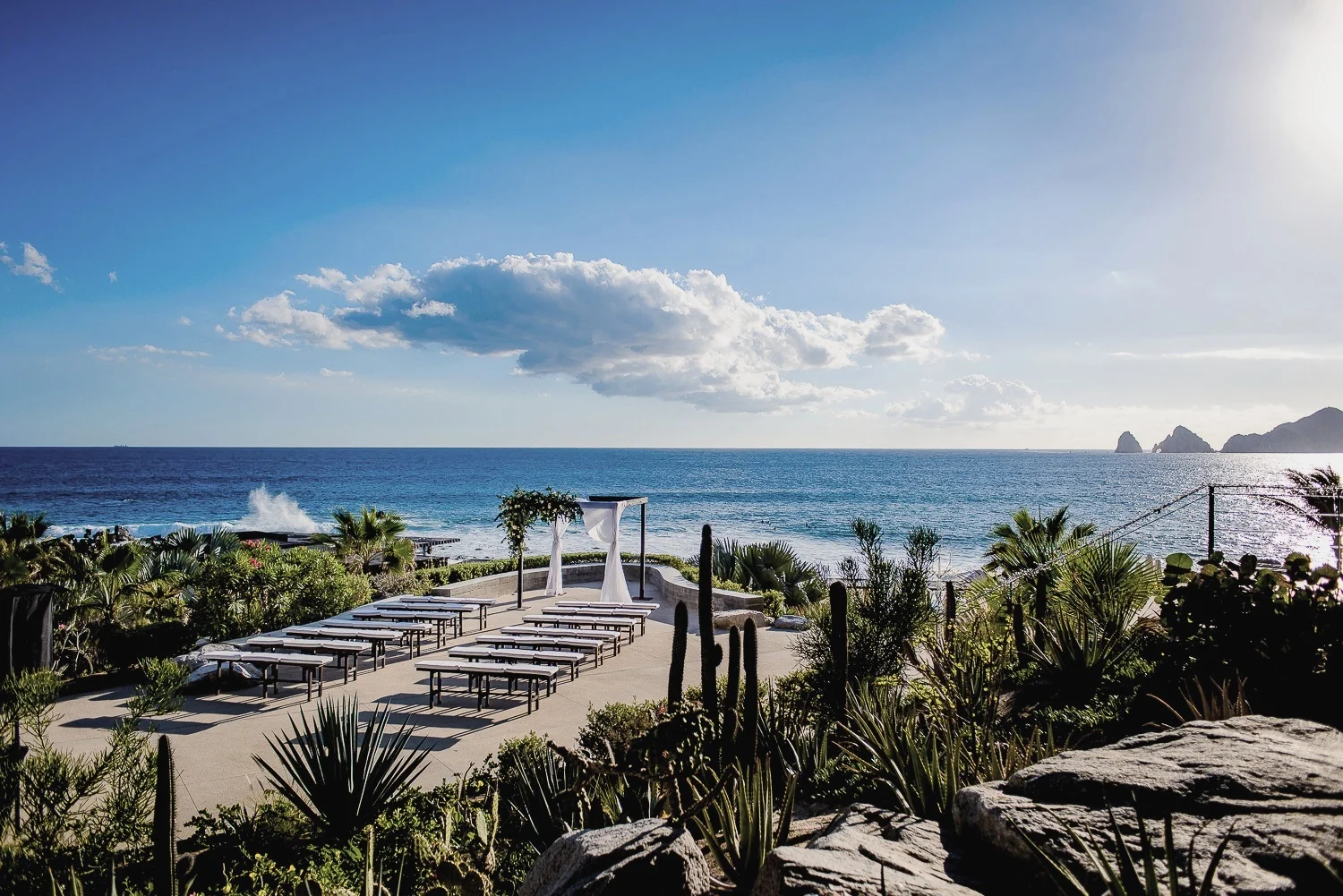 Simple wooden ceremony canopy. Framed by soft gauzy drapes billowing in the breeze and a romantic display of lush white garden roses. Simple adornment for an already spectacular setting! Low upholstered wooden benches keep the Ocean view and the Arch Rock formation uninterrupted.