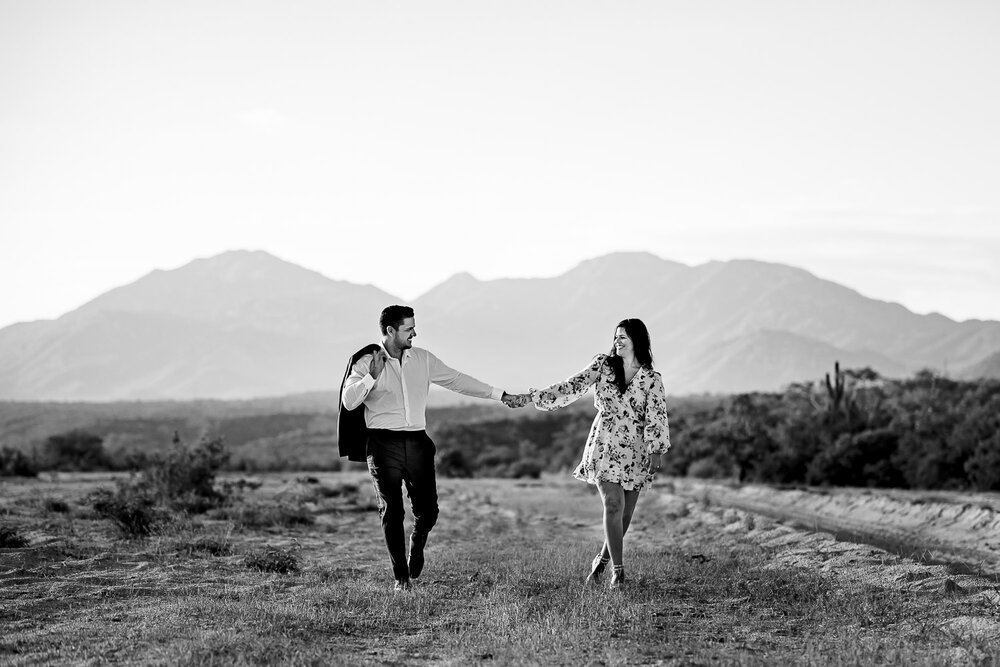 Couple walking and having fun, beautiful mountains are behind them, during their engagement photos in Cabo San Lucas
