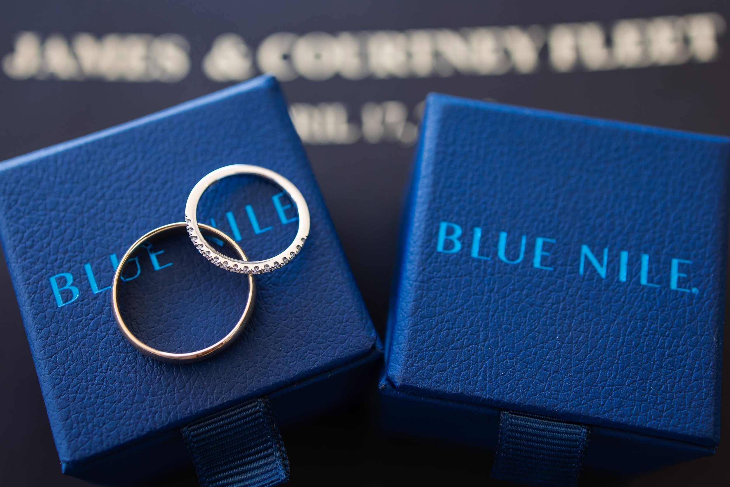  Two engagement rings on top of blue nile ring boxes with personalized text "james &amp; courtney fleet" printed in the background. one ring is a plain gold band and the other is adorned with diamonds. 