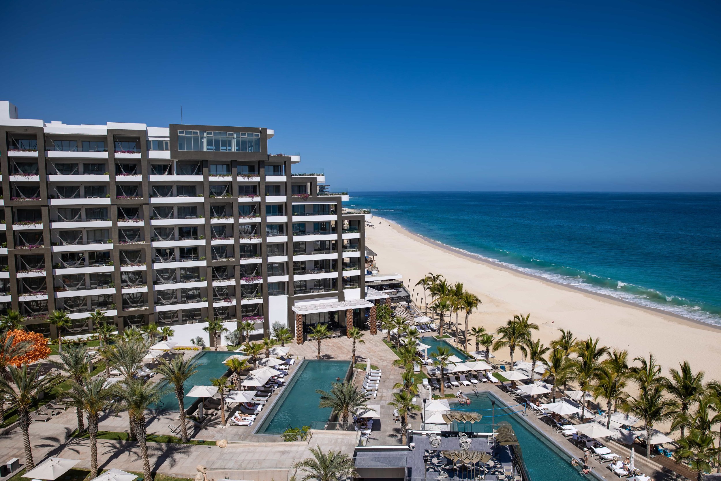  A luxurious Cabo beachfront hotel with multiple balconies overlooking a pool area surrounded by palm trees, with a wide, sandy beach and the ocean extending into the horizon. Garza Blanca 