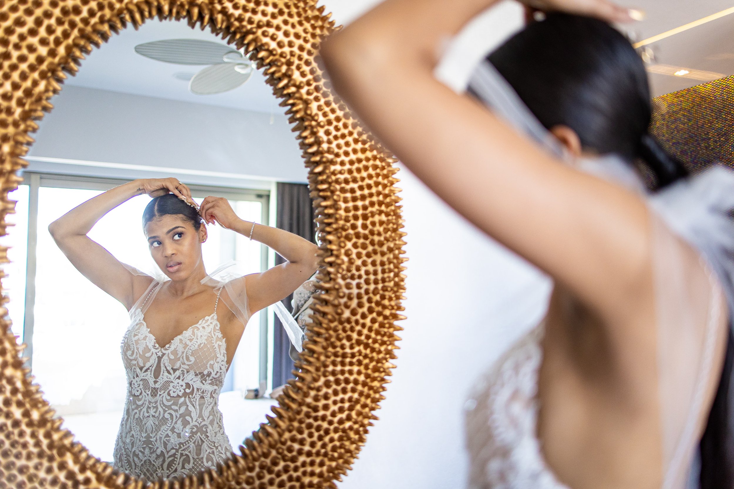  A woman in a detailed lace wedding gown adjusts her hair while looking into a round, ornate mirror in a brightly lit room. 