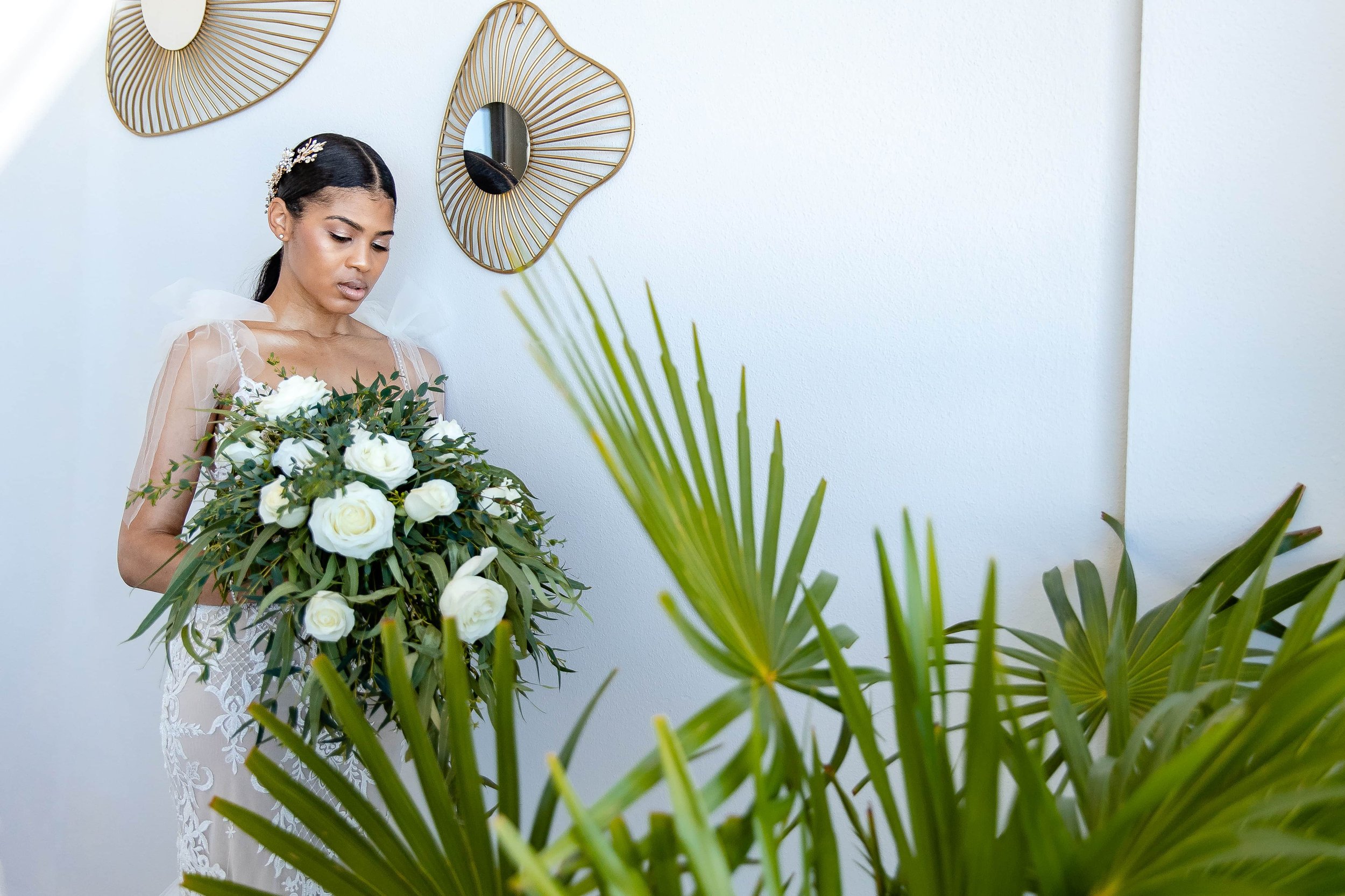  A bride in a lace gown and veil holds a large bouquet of white flowers, standing beside decorative plants and wall-mounted abstract art. 
