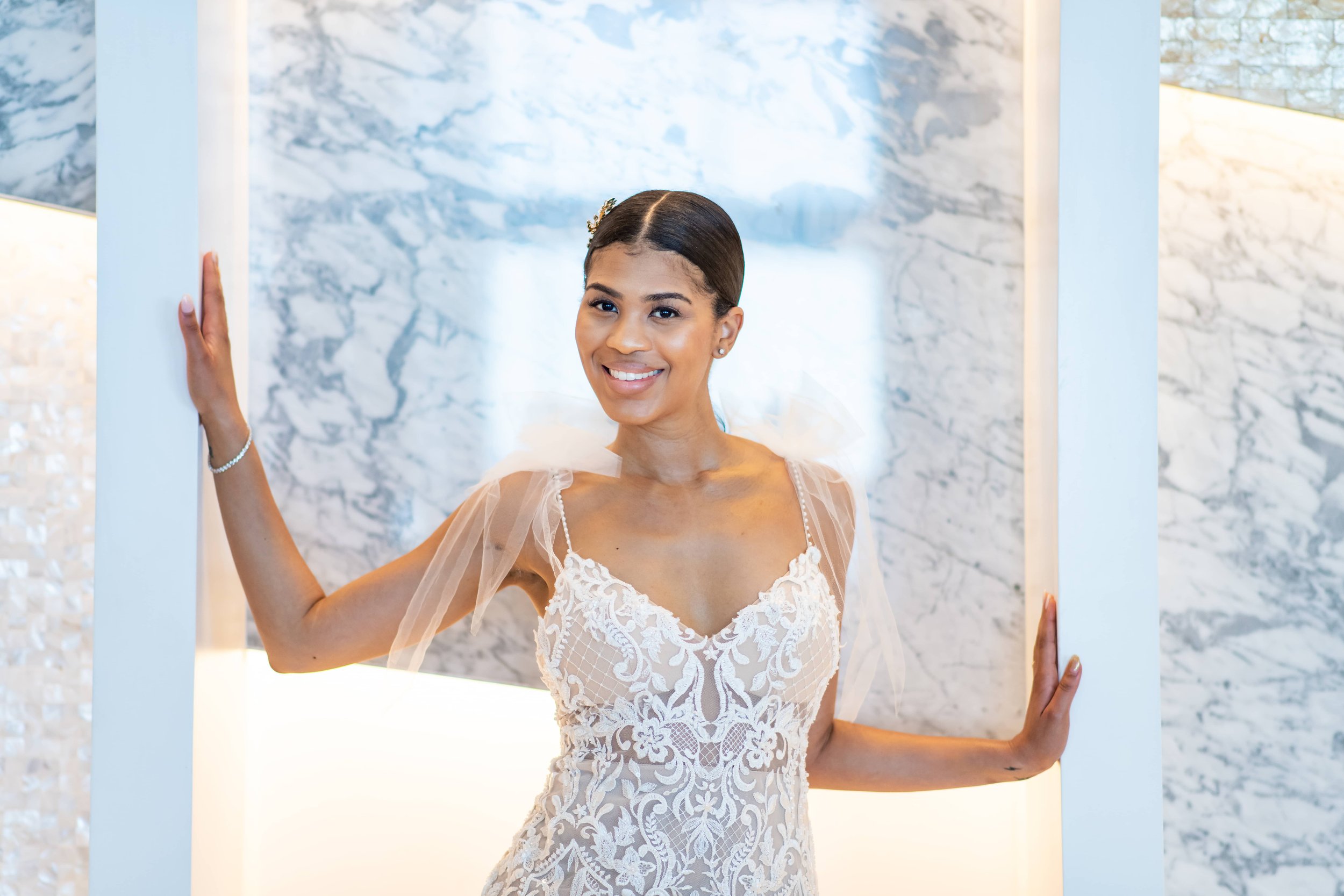  A joyful bride in a lace wedding dress with sheer sleeves poses between white columns with a marble backdrop. she is smiling broadly at the camera. 