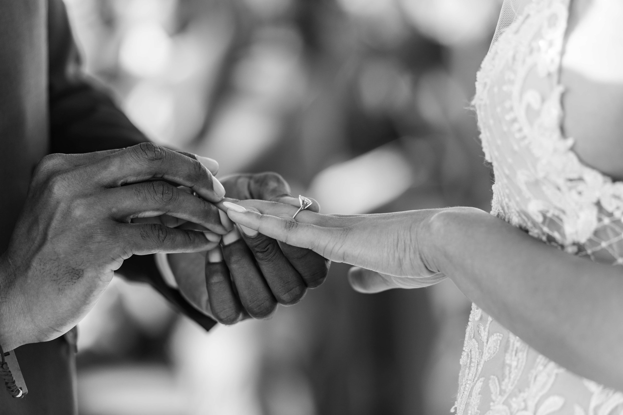  A black-and-white photo capturing a wedding ceremony moment where a groom slides a ring onto the bride's finger with both hands gently touching. 