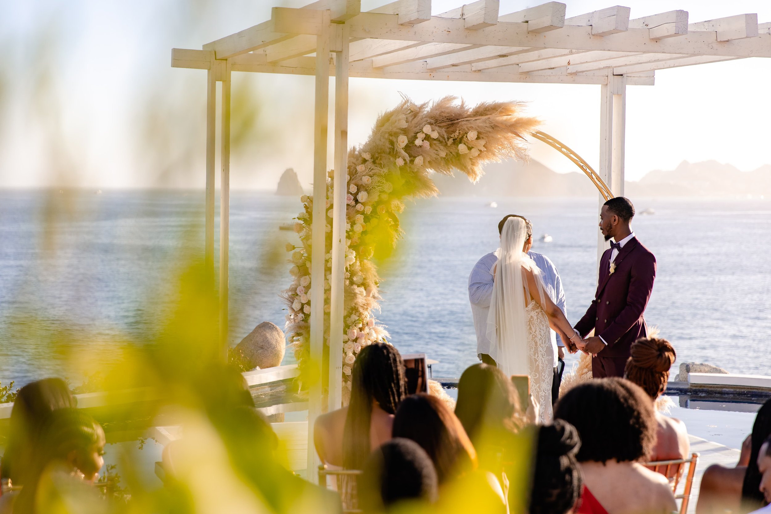  A wedding ceremony beside a tranquil sea, with the couple standing under a floral decorated archway, surrounded by guests, with Cabo Sunset Monalisa views beyond. 