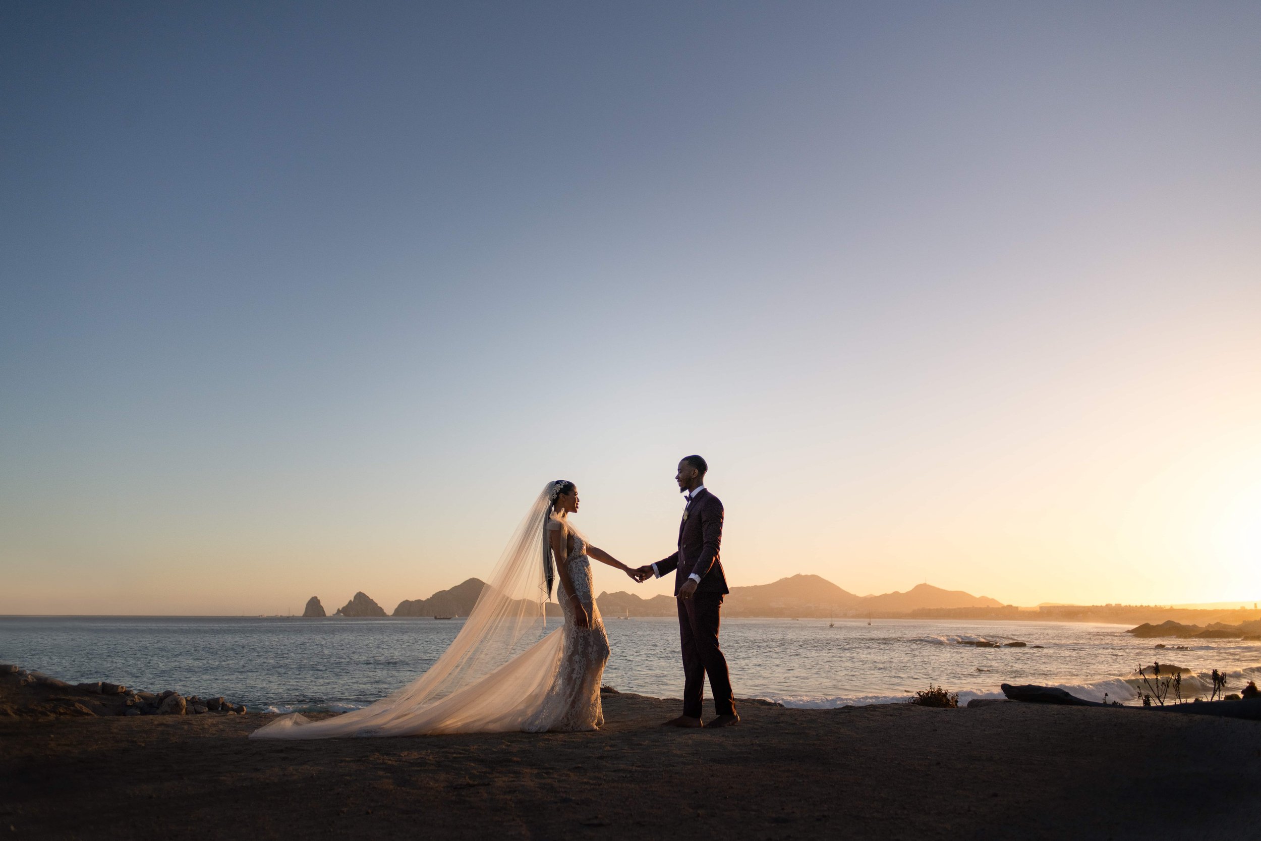  A bride and groom stand holding hands on a beach at sunset, with the bride's long veil flowing in the breeze and distant mountains and ocean in the background. 