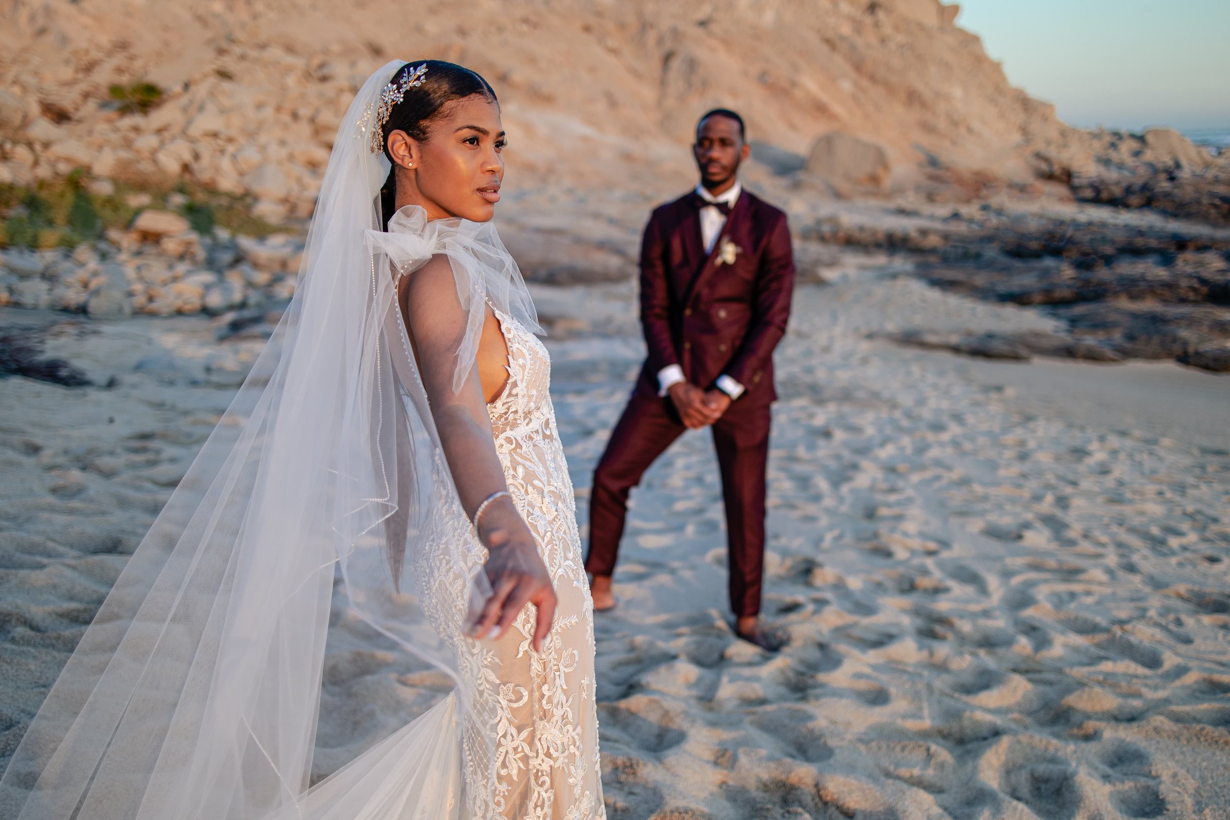  A bride in an intricate lace gown and long veil stands on a sandy beach, looking off to the side, with a groom in a sharp brown suit looking towards her in the background by rocky terrain. 