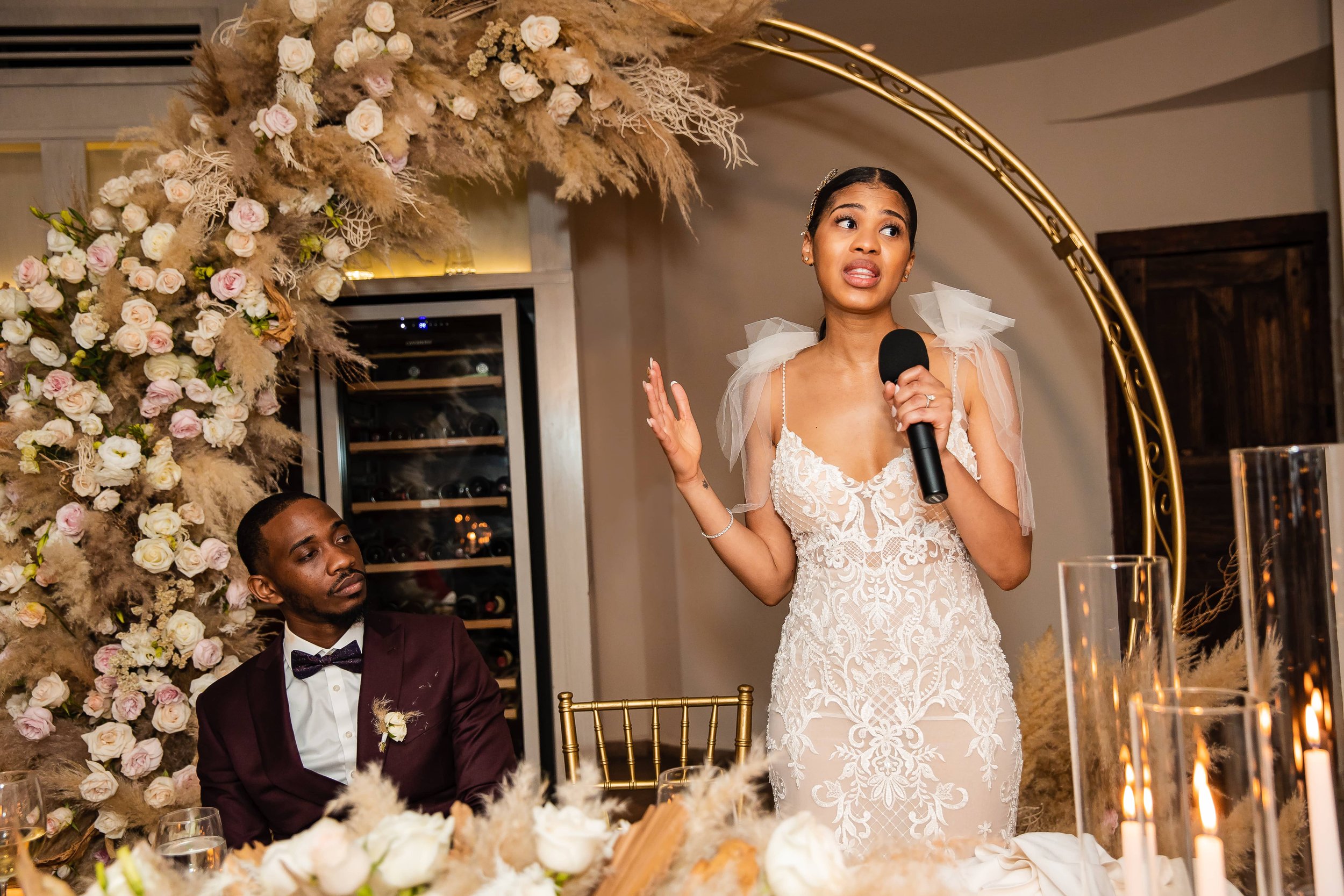  A bride in a white gown speaks into a microphone at a wedding reception, gesturing with her hands, as the groom looks on seated next to her under a floral arch. 