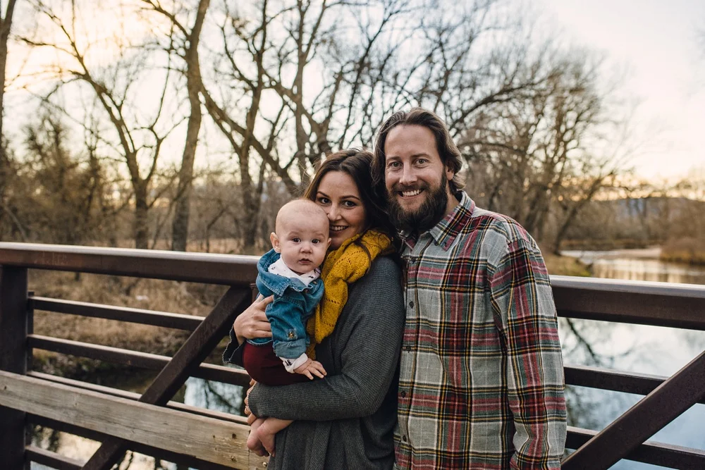A young family on a bridge over the Poudre River on a warm late fall evening at Legacy Park in Fort Collins, Colorado. Family portrait photography by Sonja Salzburg of Sonja K Photography.&nbsp;