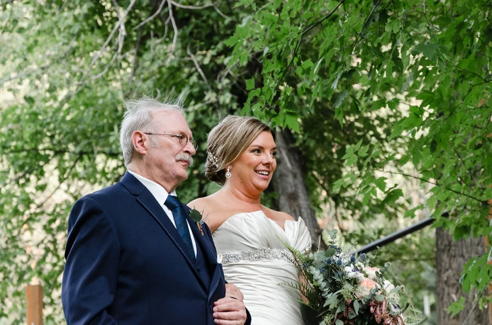 A bride and her father walk down the aisle together at a wedding at Wedgewood Weddings on Boulder Creek outside of Boulder, Colorado. Wedding portrait photography by Sonja Salzburg of Sonja K Photography.