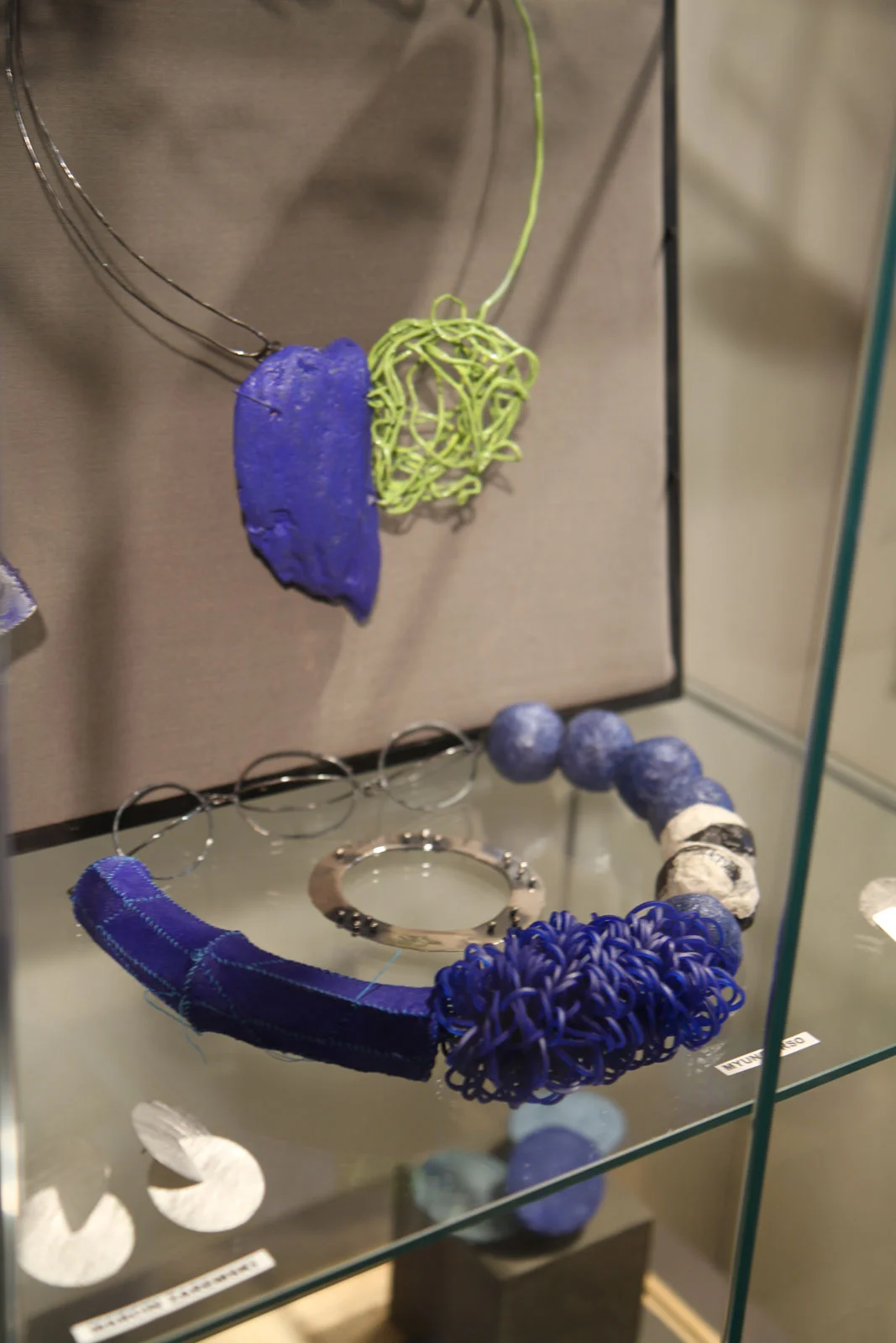 Myung Urso's multimedia necklaces, incorporating some sort of rubber/plastic and traditional handmade paper, or hanji as it is called in Korea, with acrylic paint. The piece behind it may be using wood for the broad rectangular shape.