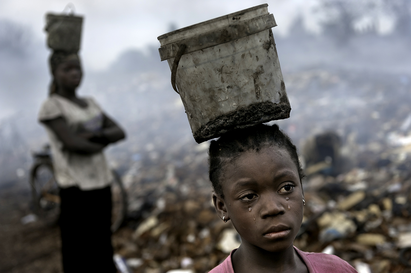 In Ghana in an e-waste dump that kills nearly everything that it touches, Fati, 8, works with other children searching through hazardous waste in hopes of finding whatever she can to exchange for pennies in order to survive. While balancing a bucket on her head with the little metal she has found, tears stream down her face as the result of the pain that comes with the malaria she contracted some years ago. This is work she must do to survive. © Renée C. Byer