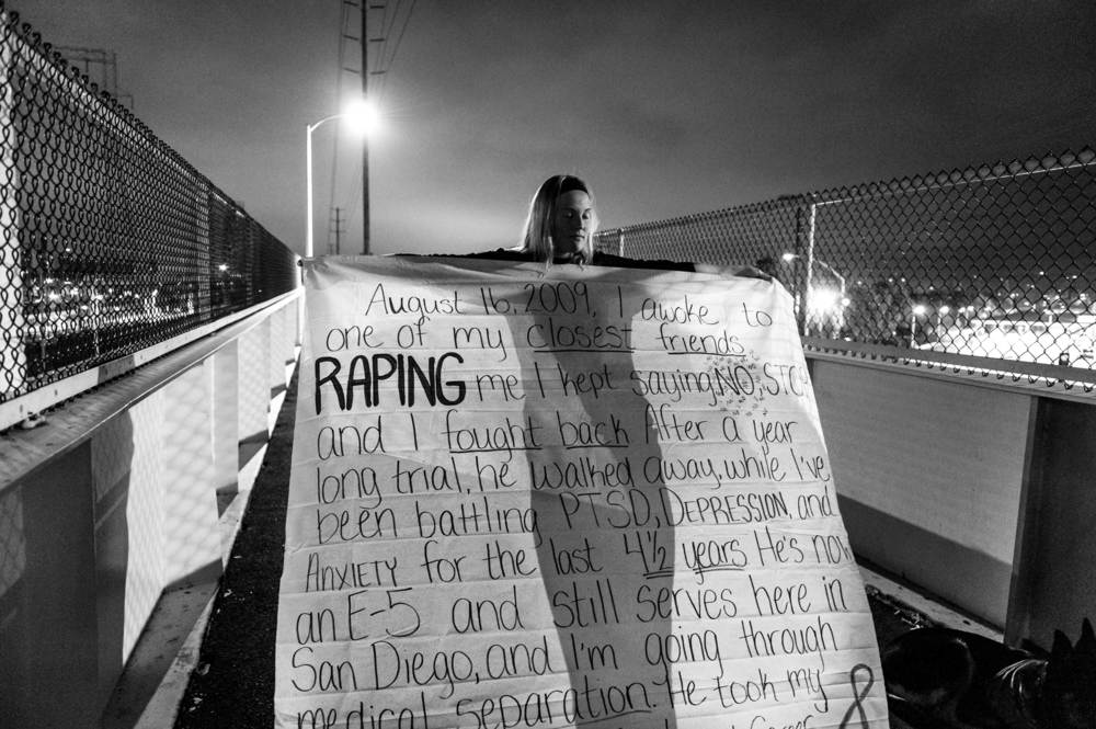   The War Within: Melissa Bania holds her banner, before hanging it on the foot bridge across from the entrance to Naval Station San Diego. US Navy Military Sexual Trauma survivors got together to make banners inscribed with their sexual assault expe