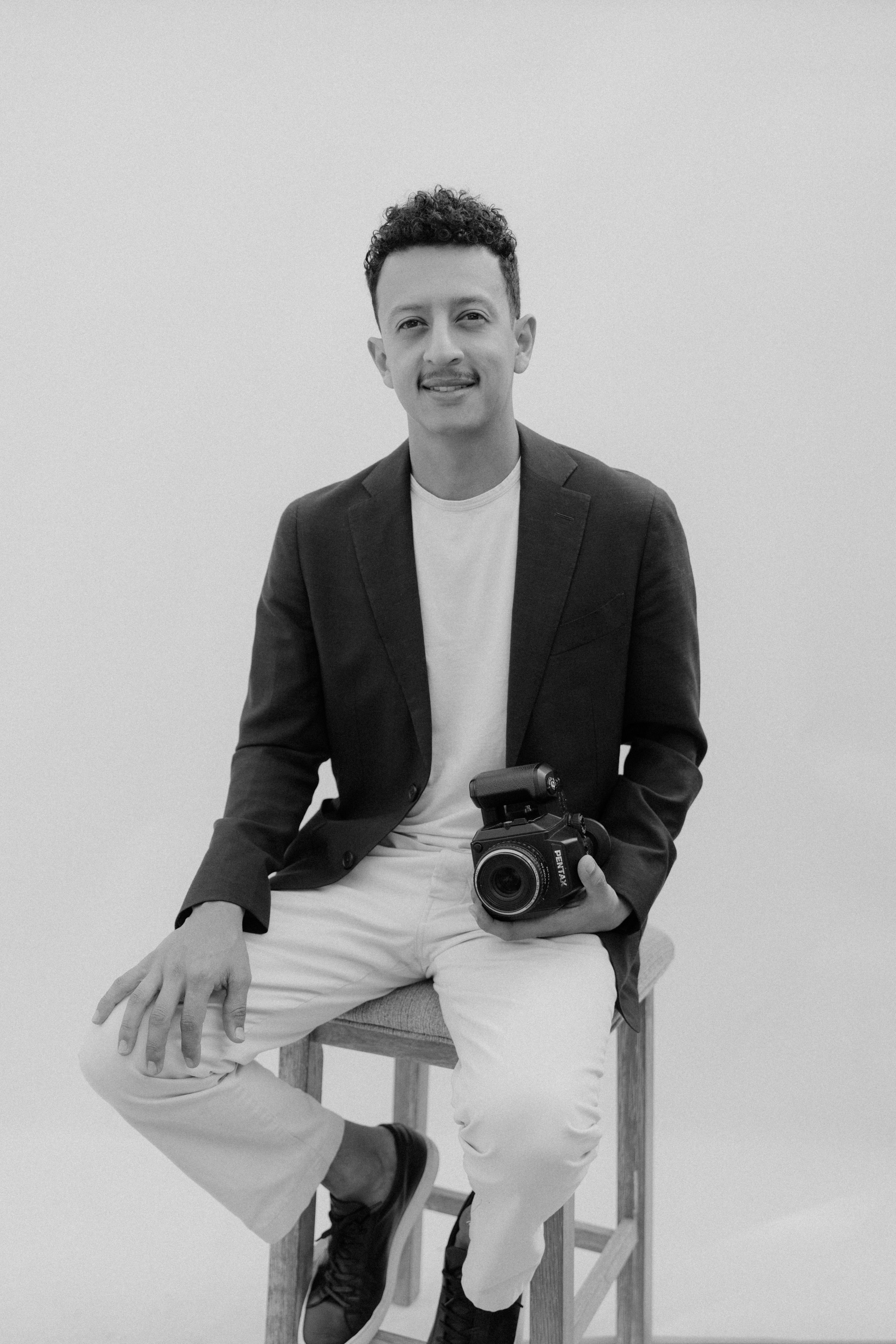 Man sitting on a stool, holding a camera, wearing a blazer, white shirt, and white pants, in a minimalist setting.
