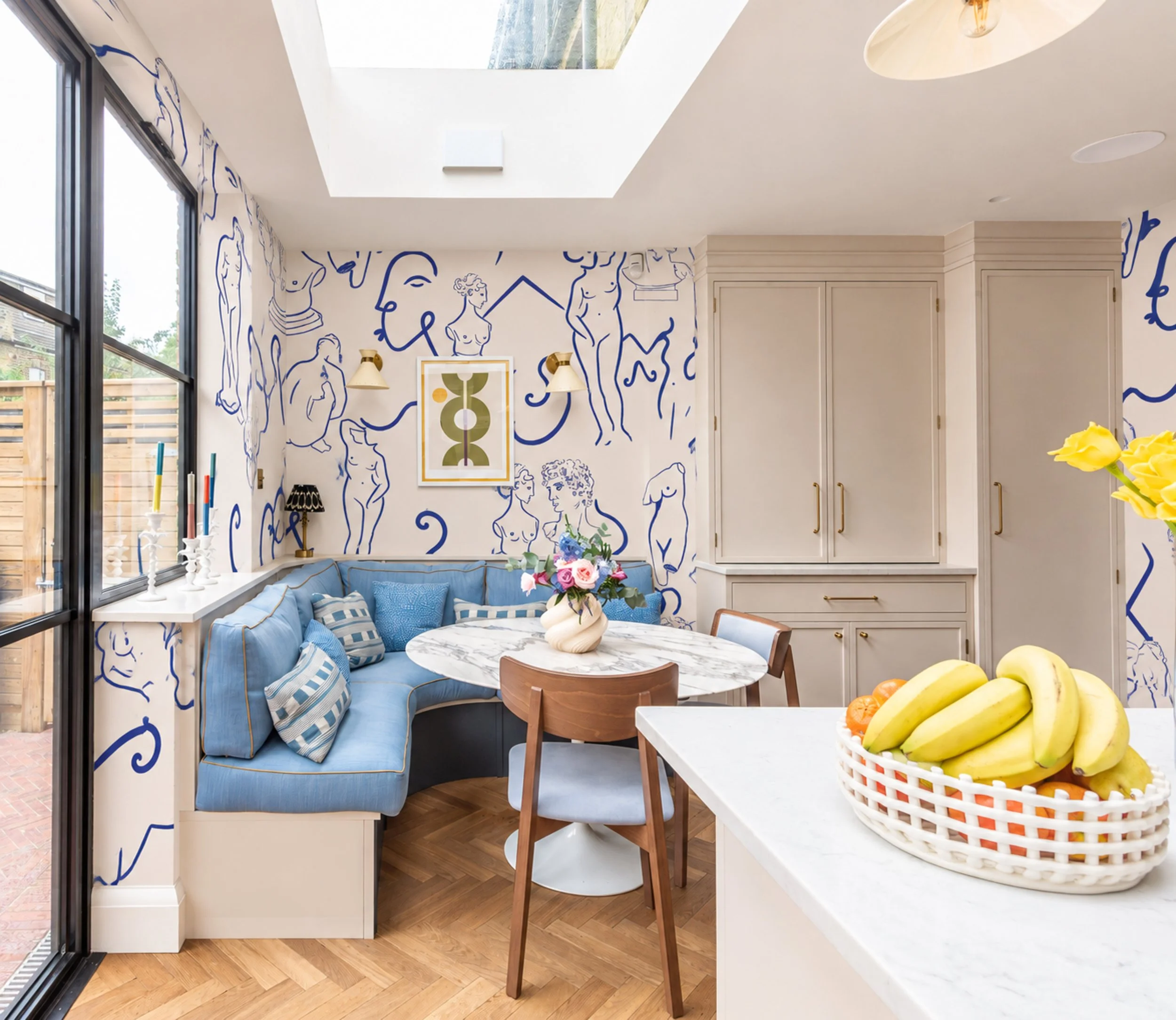 A cozy kitchen corner with a marble table, a light blue cushioned window seat with striped pillows, a basket of bananas, and abstract line art on the walls featuring human figures and faces, with a ceiling skylight letting in natural light.