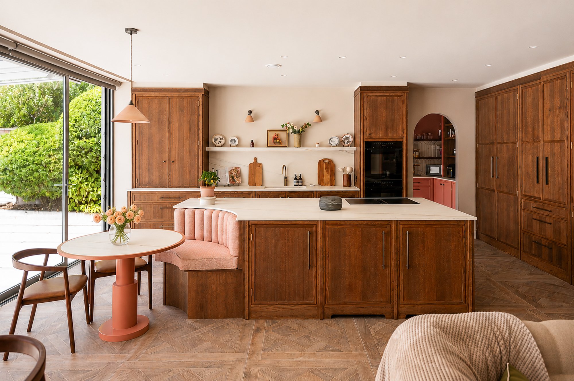 Modern kitchen with wooden cabinets, a white marble island, pink dining chair, round pink table with a vase of pink roses, and sliding glass doors overlooking greenery.