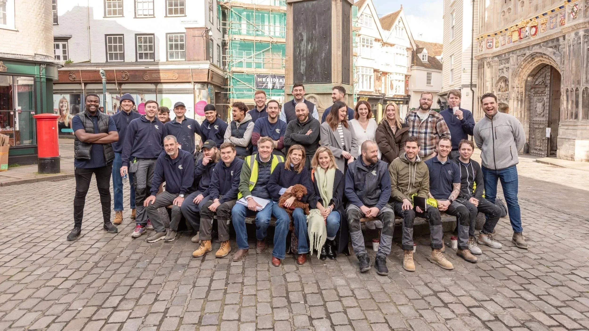 A large group of people, including men, women, and children, gathered outdoors in front of historic buildings with cobblestone pavement. They are smiling and posing for the photo, some wearing casual clothes, others in uniform or work clothes. There is a dog in the front. The background includes scaffolding on one building and detailed architecture.