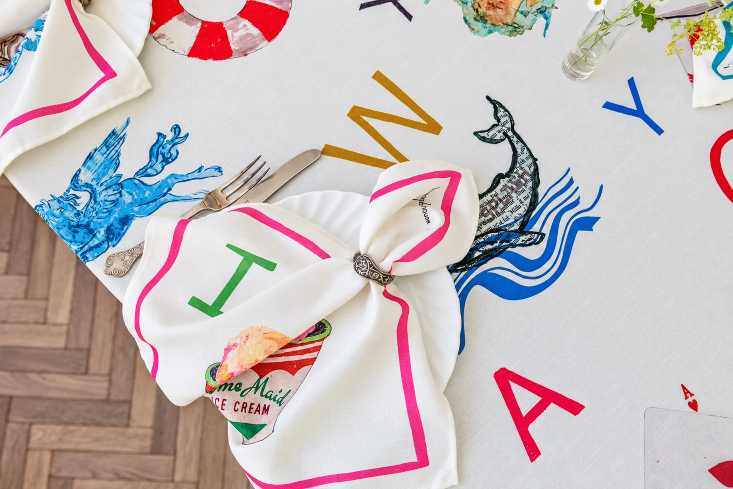 A table covered with a white cloth featuring colorful text and illustrations. In the foreground, there's a white napkin with pink and green accents folded into a decorative shape, with silverware placed on it. Several other decorated napkins, a small bouquet of flowers in a clear glass vase, and a scoop of colorful ice cream are also visible on the table.