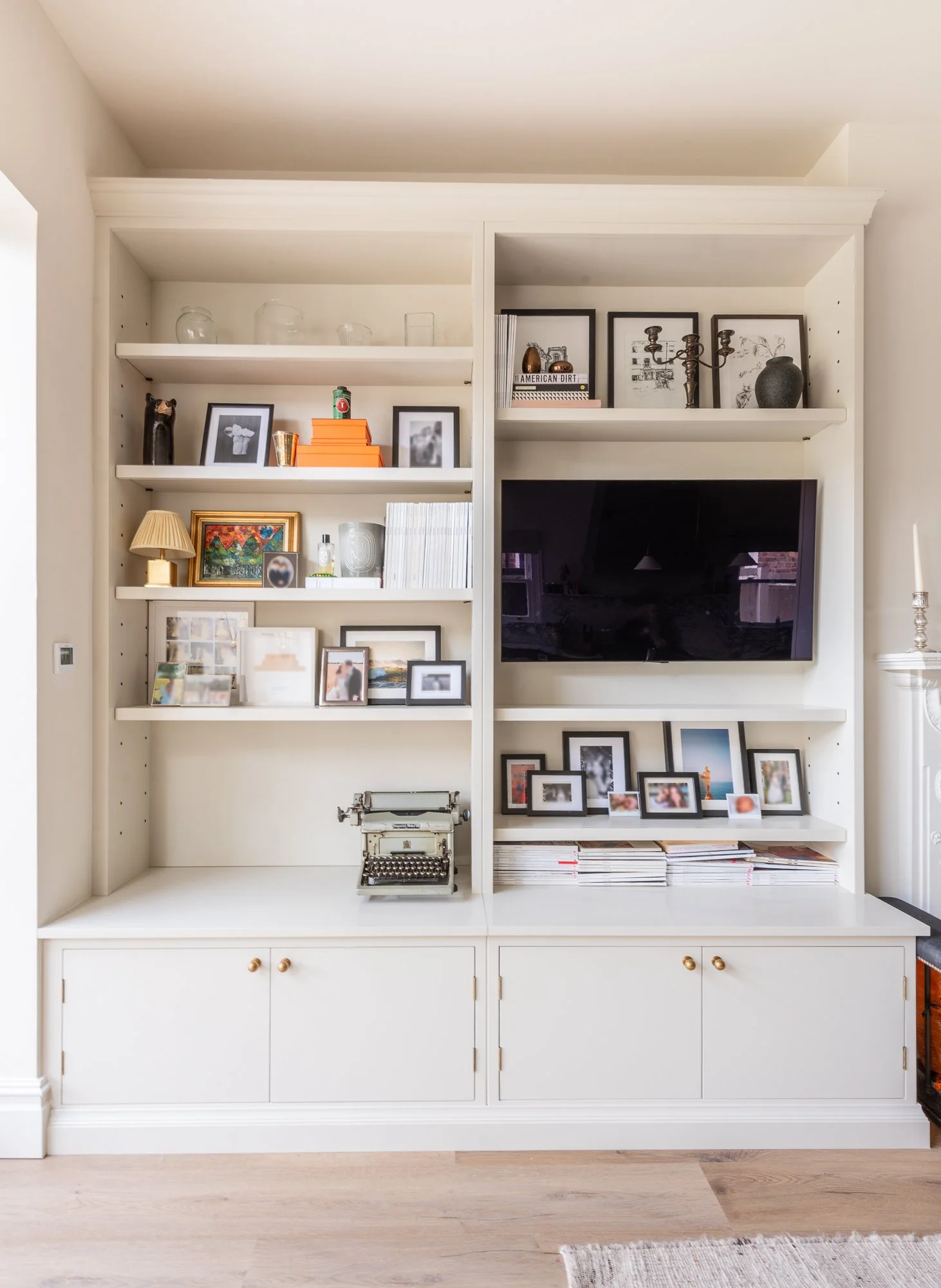 White built-in wall shelf with decorative items, framed photos, a vintage typewriter, and a mounted flat-screen TV in a living room.