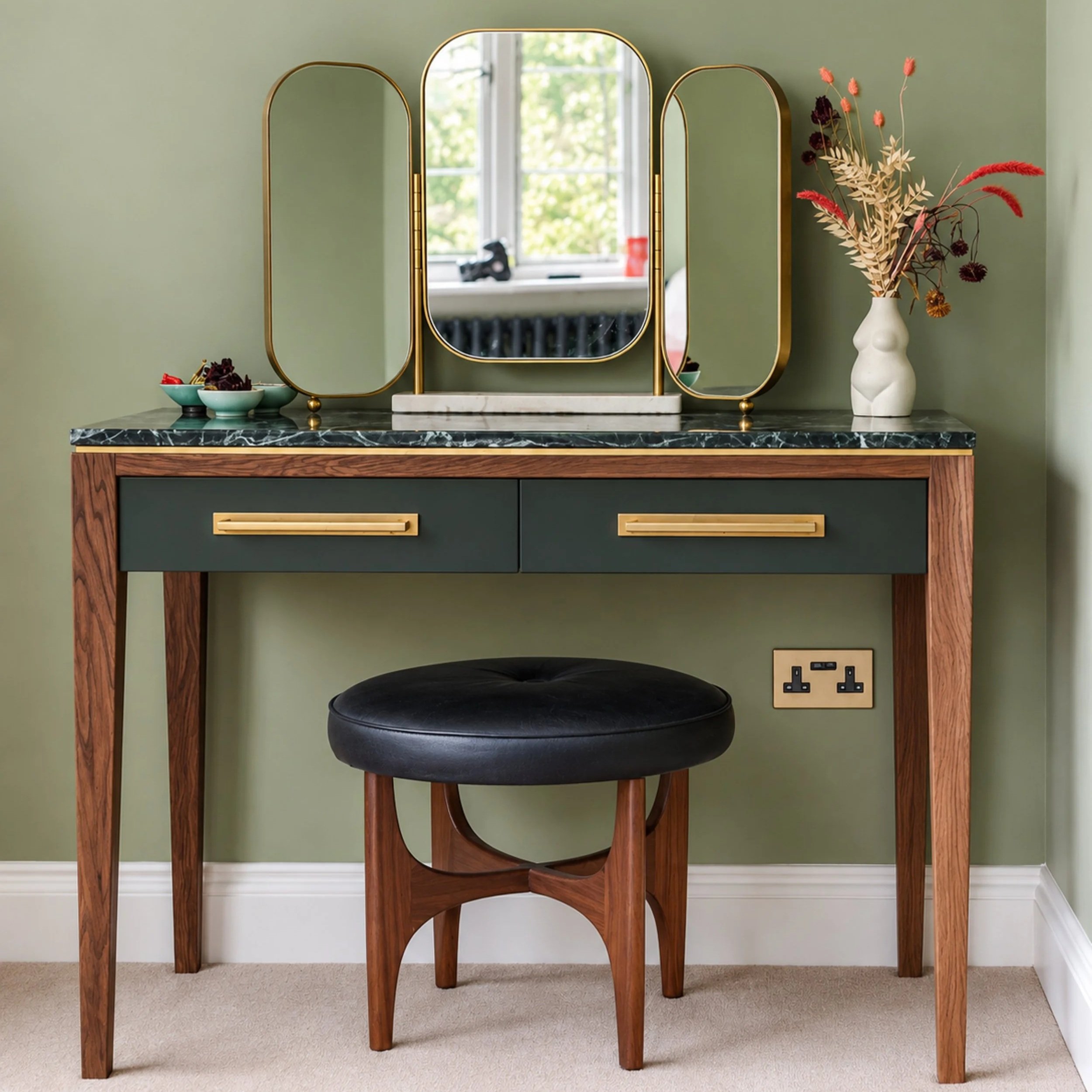 A black chair with a rounded seat and backrest in front of a green wall, positioned under a wooden console table with a dark marble top, with a beige carpeted floor and a white baseboard. To the right, a white vase with a human torso shape contains a bouquet of dried pink, dark purple, and beige foliage. Three small bowls with snacks are on the left side of the table, and a electrical outlet is visible on the wall near the floor.