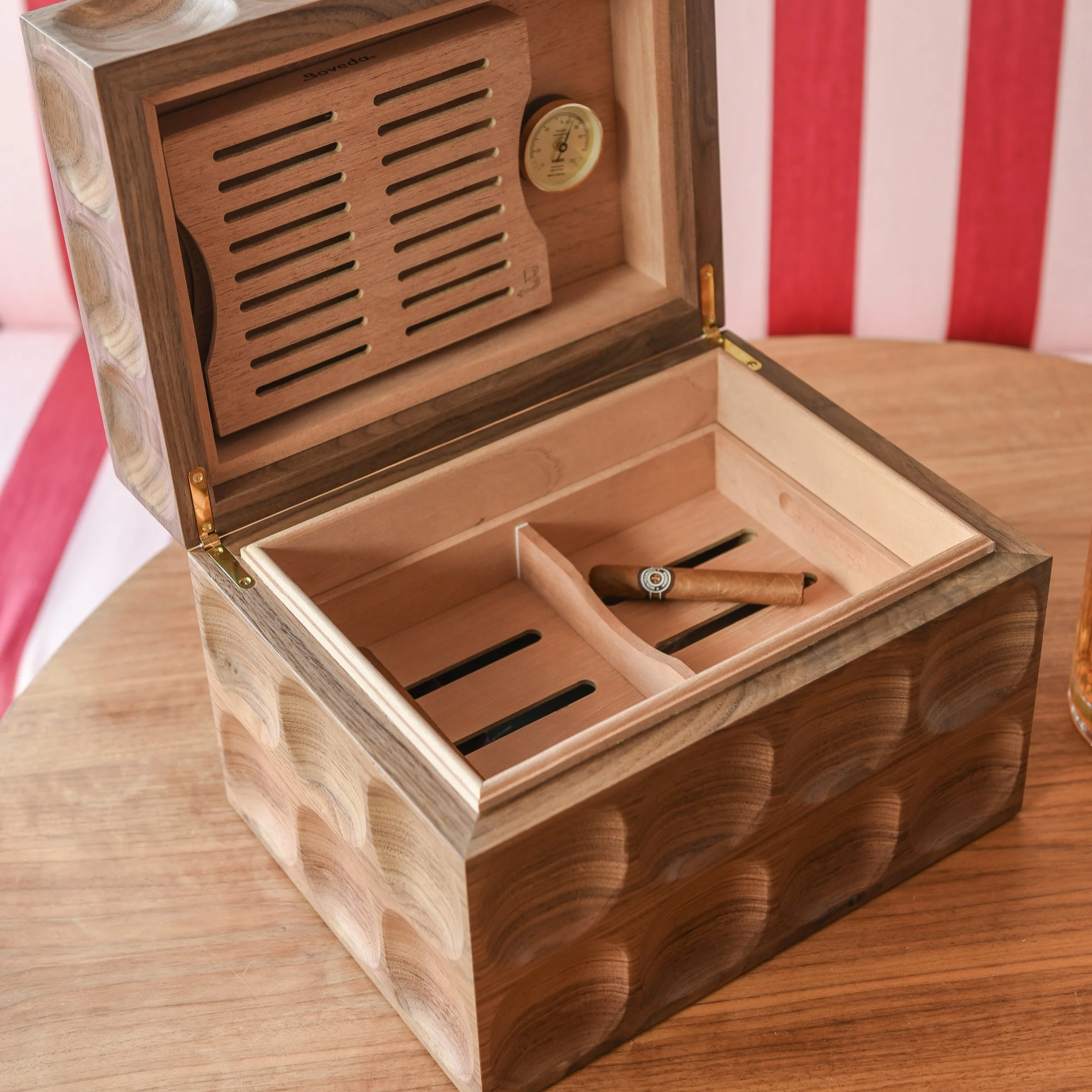 An open wooden cigar box containing a single cigar and a hygrometer, with a wood surface underneath and a wall with red and white vertical stripes in the background.