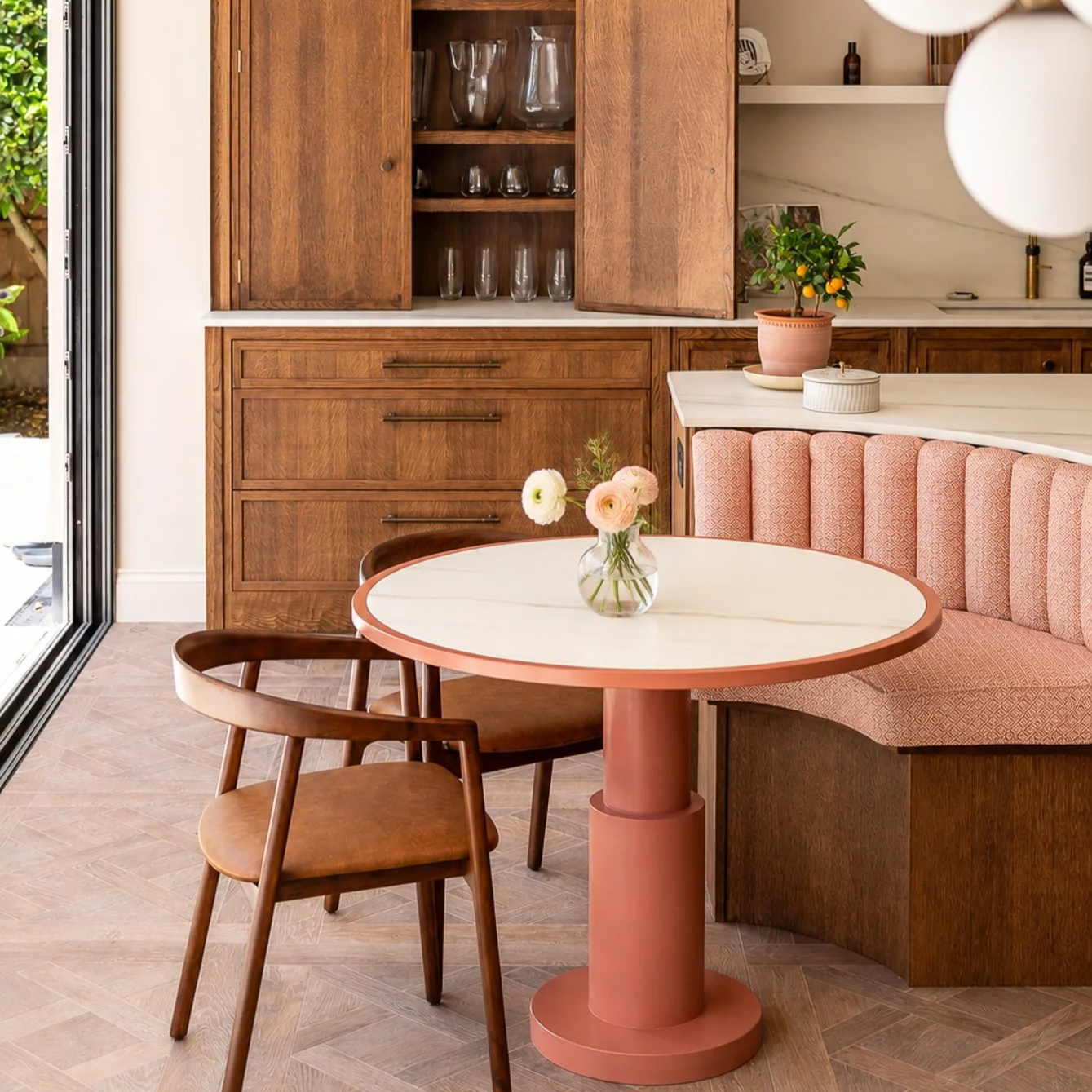 A cozy dining area with a round white table, a pink base, a glass vase with flowers, two wooden chairs, and a pink upholstered banquette. Behind is a wooden cabinet with glassware, a pink potted plant, and decorative items.