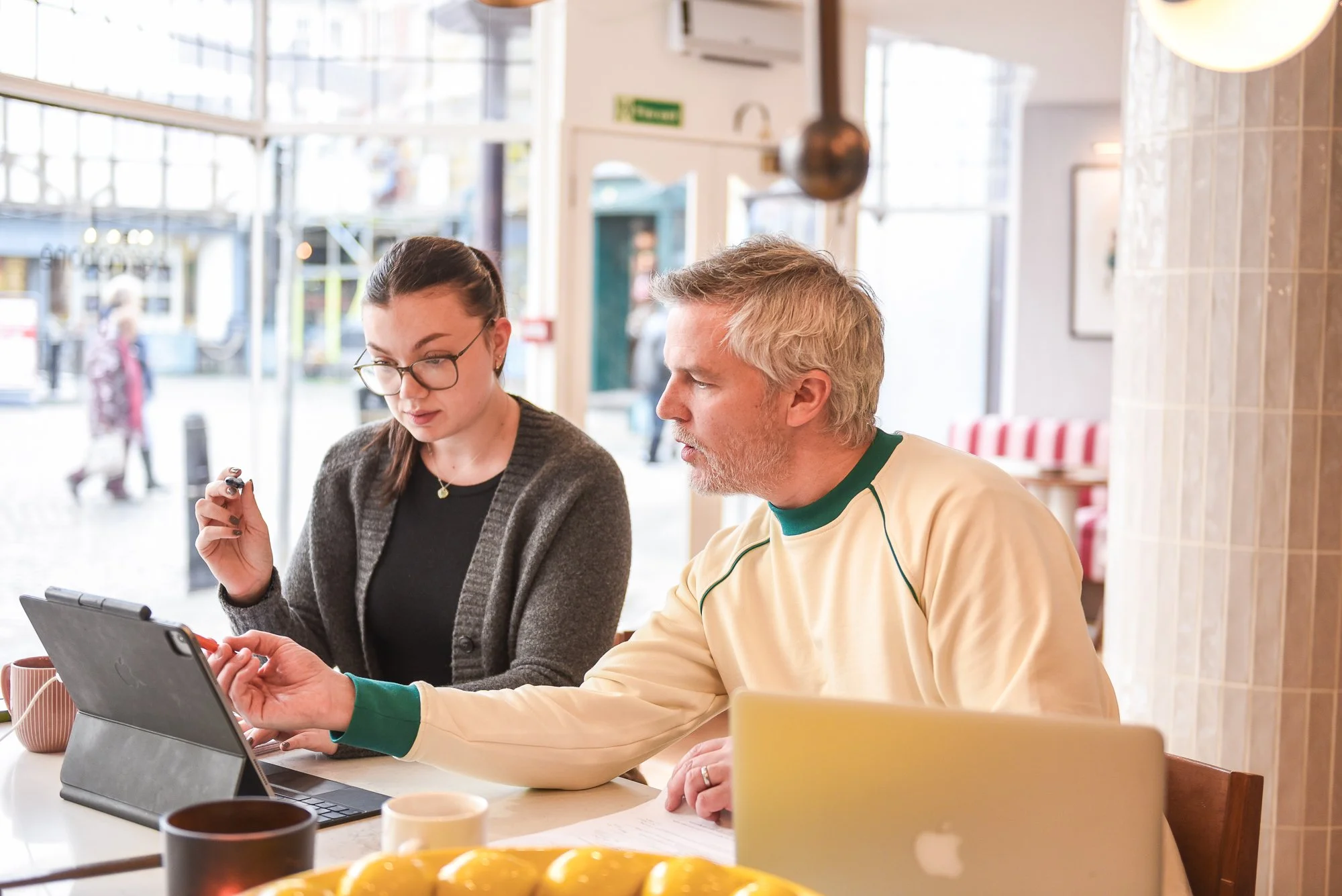 Two people sitting at a table in a cozy cafe, looking at a tablet device together. The woman on the left wears glasses and a cardigan, and the man on the right has gray hair and a beard, wearing a cream-colored sweater with green accents. There are mugs and a yellow bowl on the table, and large windows letting in natural light.