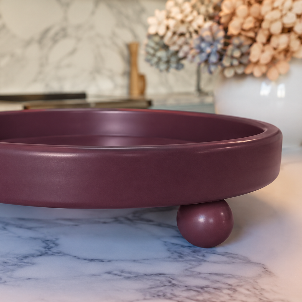A close-up of a round, maroon-colored tray or stand with a decorative spherical leg, placed on a white marble surface. In the background, there is a white vase with pink and purple hydrangea flowers and a blurred kitchen backdrop.