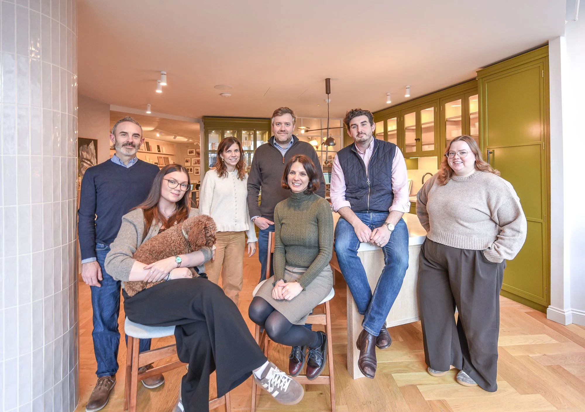 A group of seven people, including four women and three men, posing together in a cozy, modern kitchen with green cabinets and wood floors.