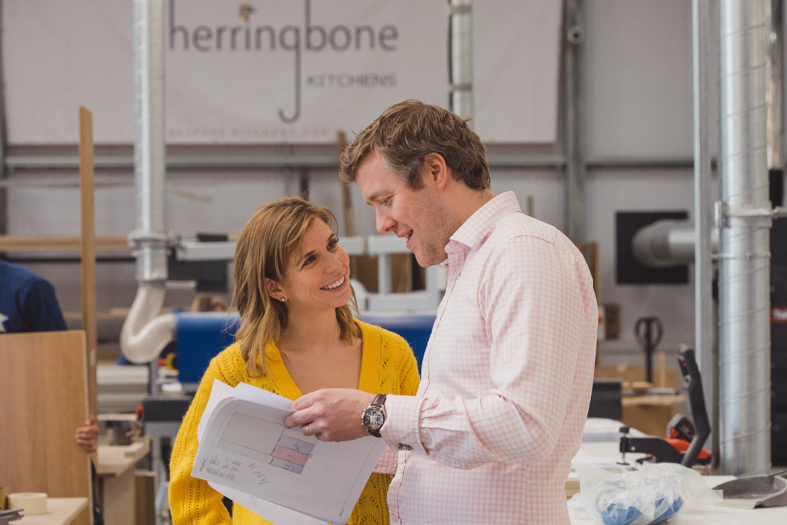 A woman in a yellow sweater and a man in a light pink shirt are smiling and looking at each other while holding a construction plan in a workshop or manufacturing setting.