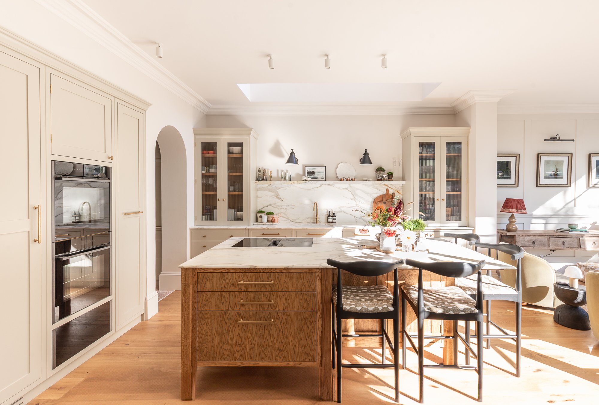 Modern kitchen with white cabinets, marble backsplash, wooden island, black bar stools, and artwork, with sunlight coming through windows.