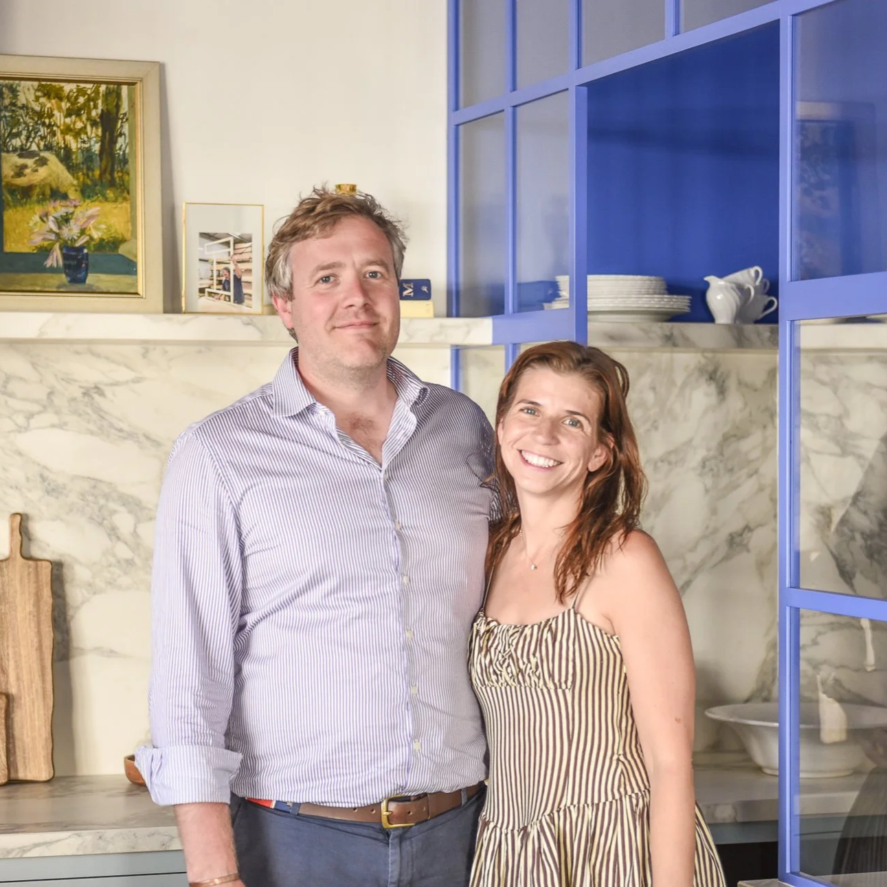 A man and woman standing in a kitchen, smiling at the camera. The man is wearing a light purple striped shirt and the woman is wearing a striped dress. The background features marble countertops, blue shelves with white dishes, and framed artwork on the wall.
