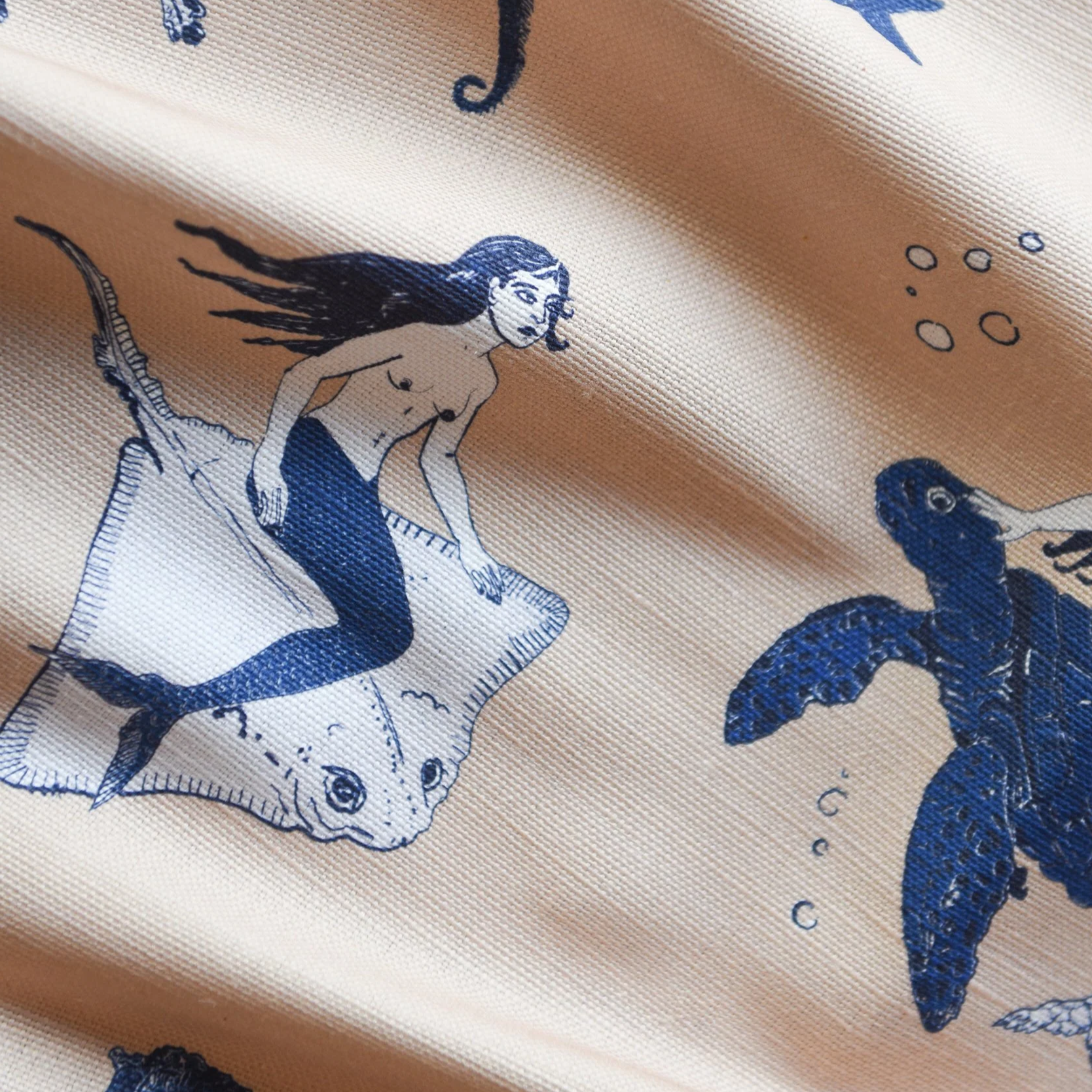 Close-up of a fabric with a printed illustration of a mermaid with long flowing hair and a fish tail, along with a cartoonish fish and bubbles.