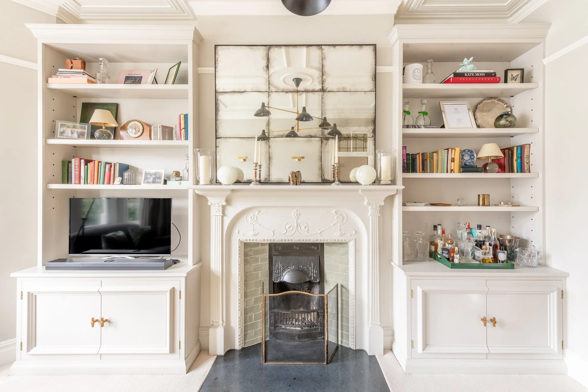 White built-in bookcases flanking a fireplace with decorative items, books, a TV, and a mirrored artwork above the fireplace