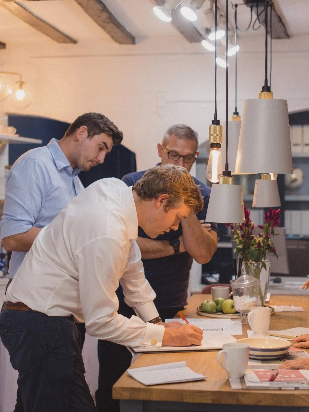 Three men in a kitchen or dining area looking at a notebook or recipe on a table, with a reference person signing or writing in the notebook. The scene has a modern decor with hanging lights, a vase with flowers, apples, and cups on the table.