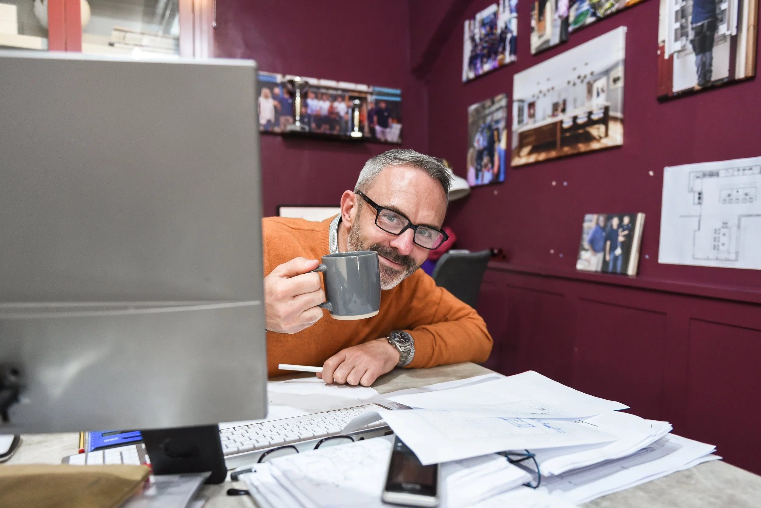 A man with glasses and a beard, wearing an orange sweater, sitting at a cluttered desk with papers, a keyboard, and a computer monitor. He is holding a gray mug and smiling at the camera. The background has maroon walls with various photos and papers pinned or taped to them.
