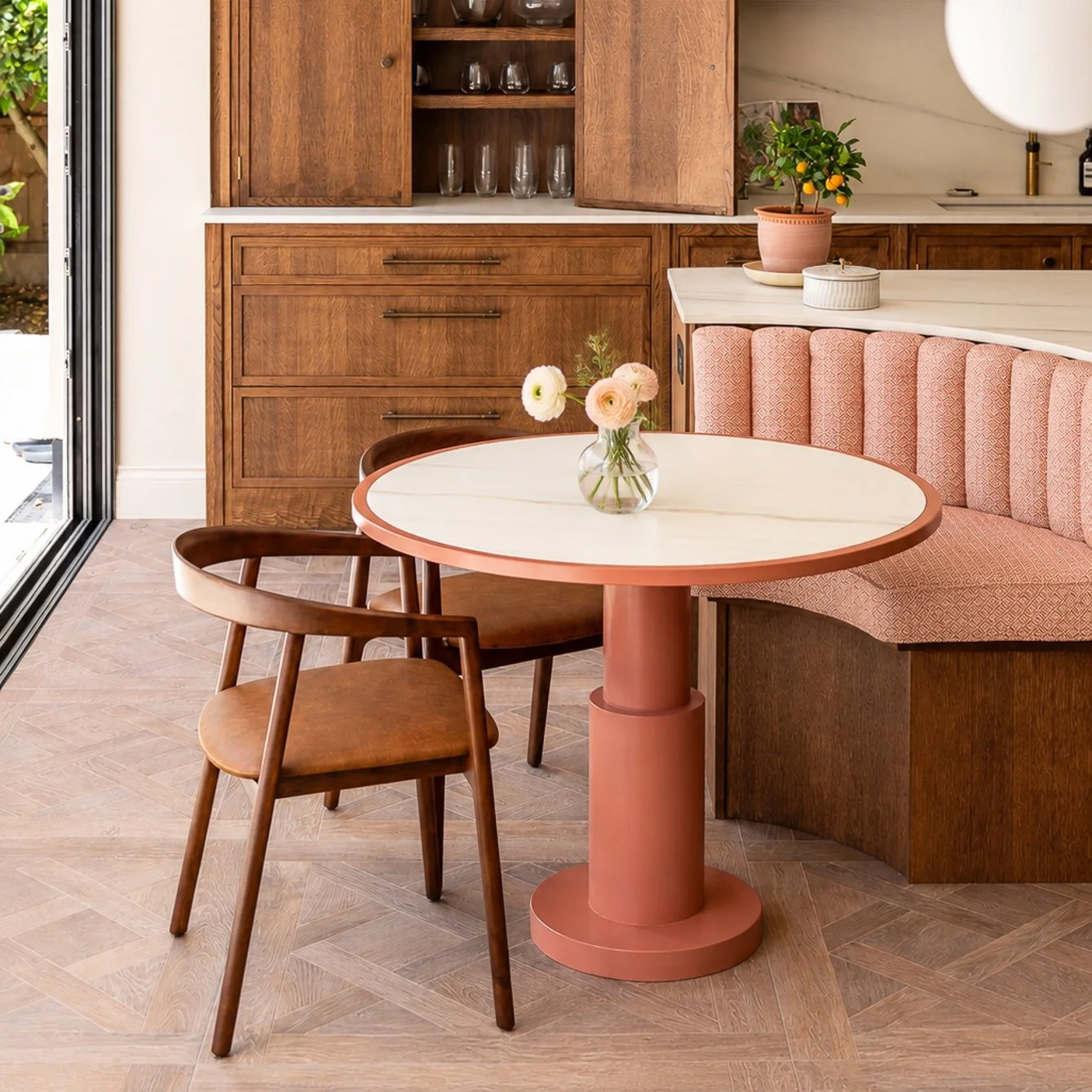 Interior view of a dining area with a round white marble table, pink pedestal base, and a glass vase with white and pink flowers. There are two wooden chairs and a pink upholstered banquette. Behind, built-in wooden cabinets with glassware inside and a potted lemon tree on the white countertop.