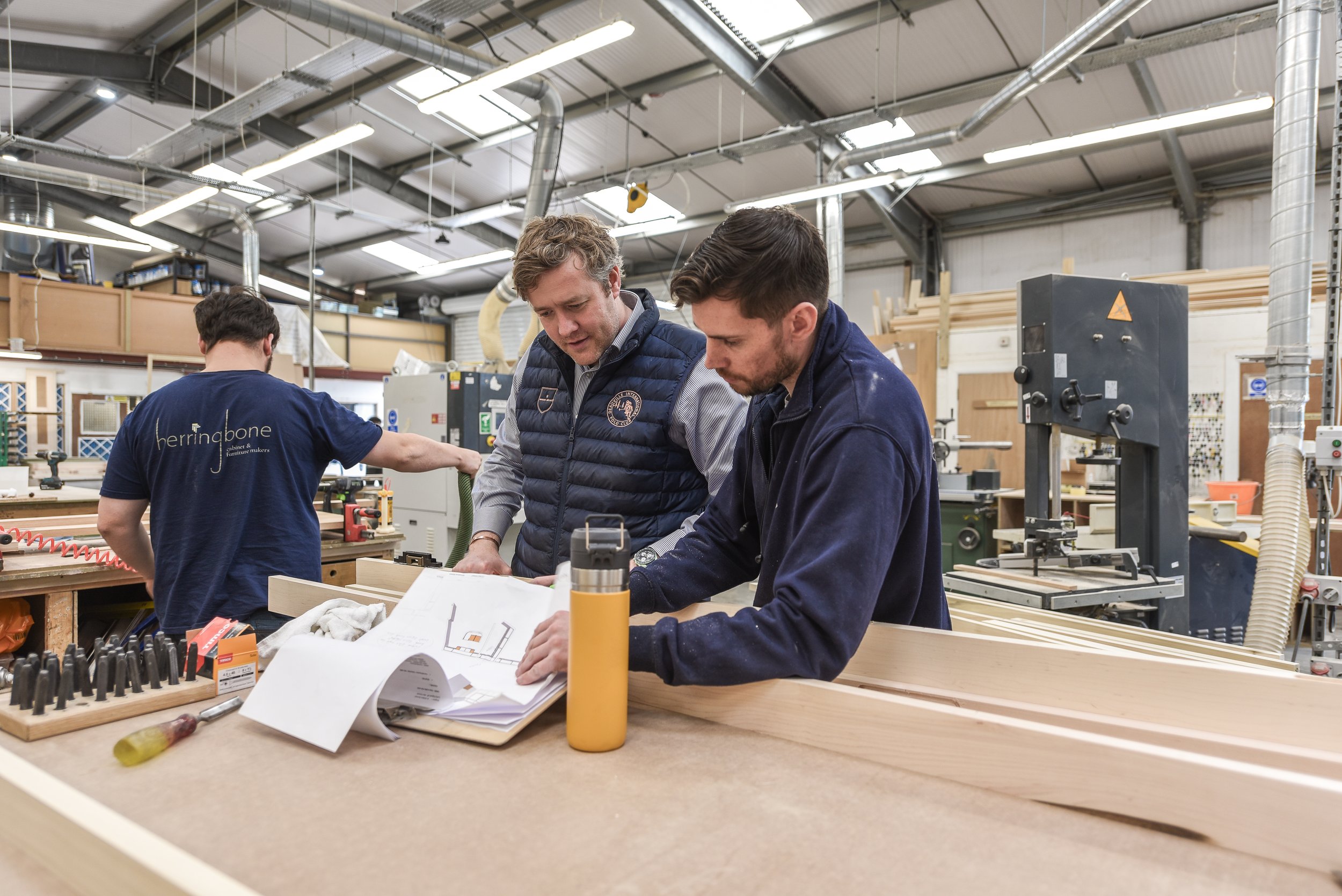 Two men and a woman in a woodworking shop examining plans or blueprints at a workbench, with woodworking tools and machines around them.