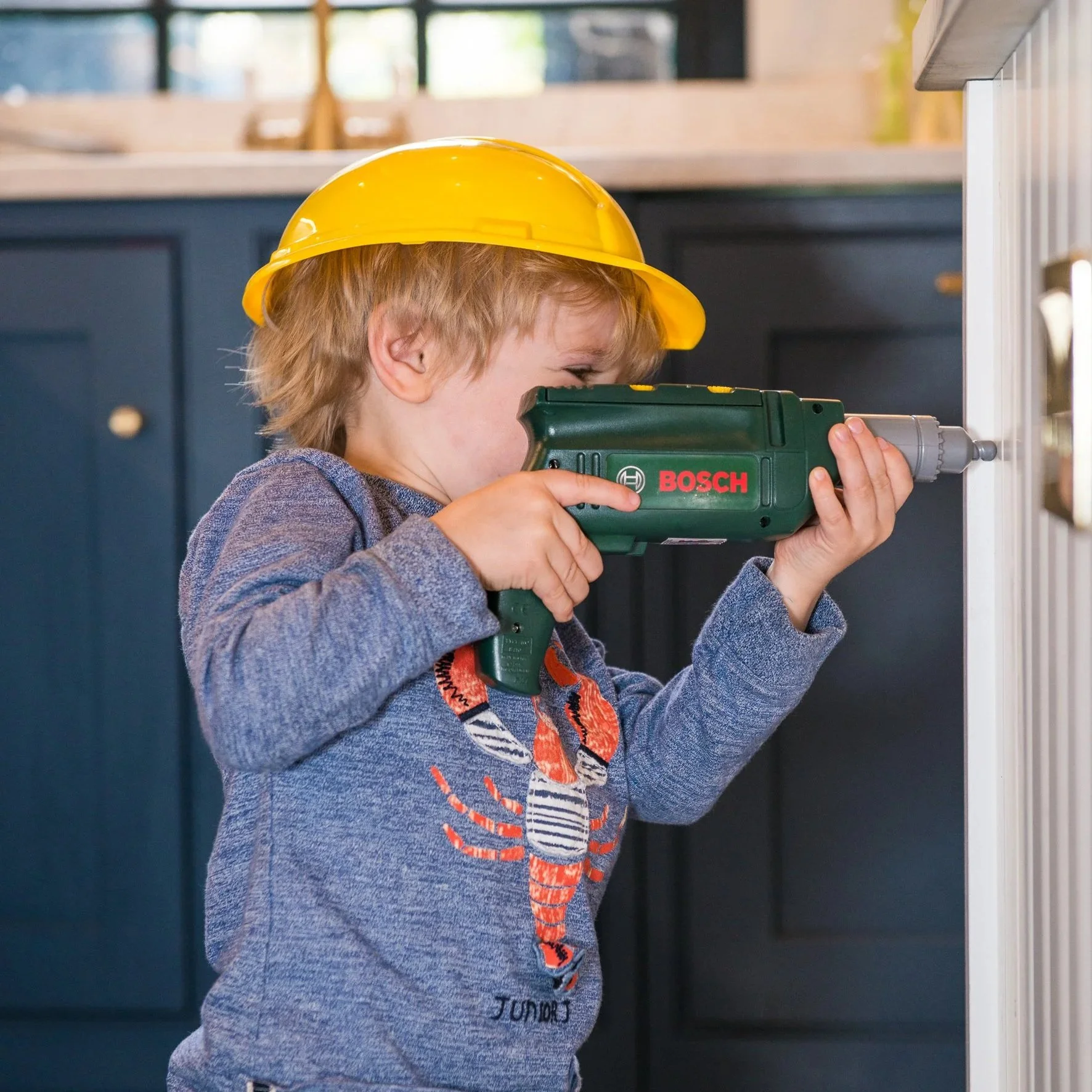 Young boy wearing a yellow construction helmet using a Bosch cordless drill to screw or drill into a surface in a kitchen or workshop.