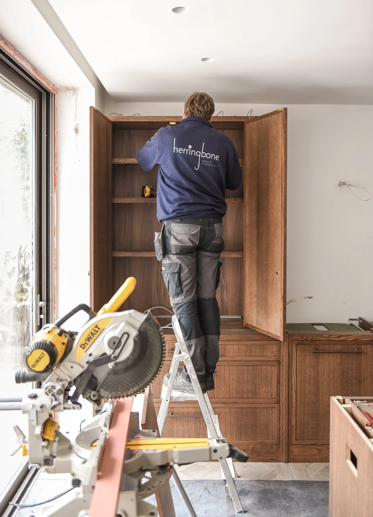 Person installing a wooden cabinet in a kitchen, using a ladder and power tools, with a table saw in the foreground, and a sliding glass door on the left.