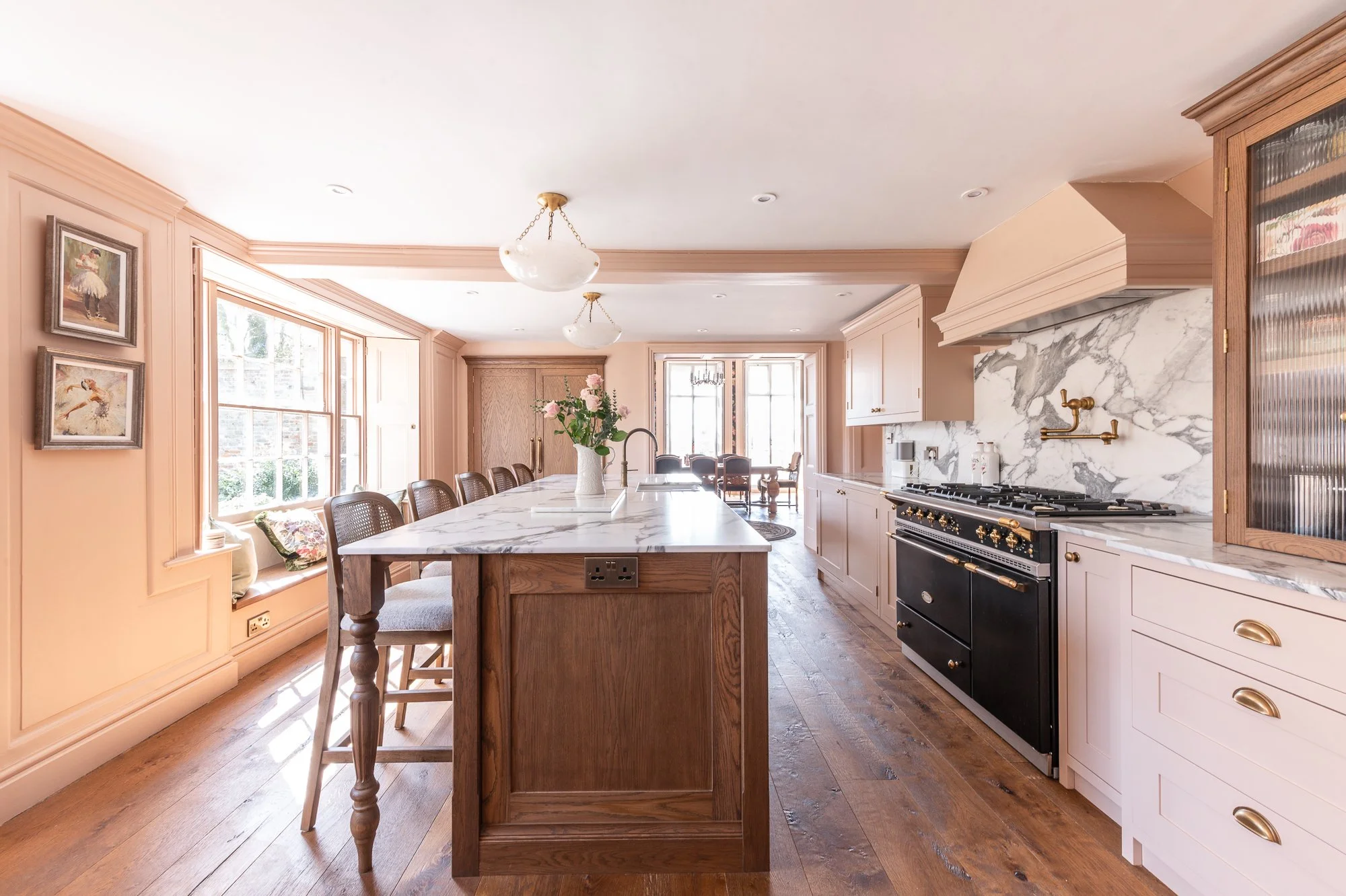 Bright kitchen with marble countertops, wooden cabinets, and pink walls, featuring a large island with a vase of pink flowers and a built-in vent above the stove.