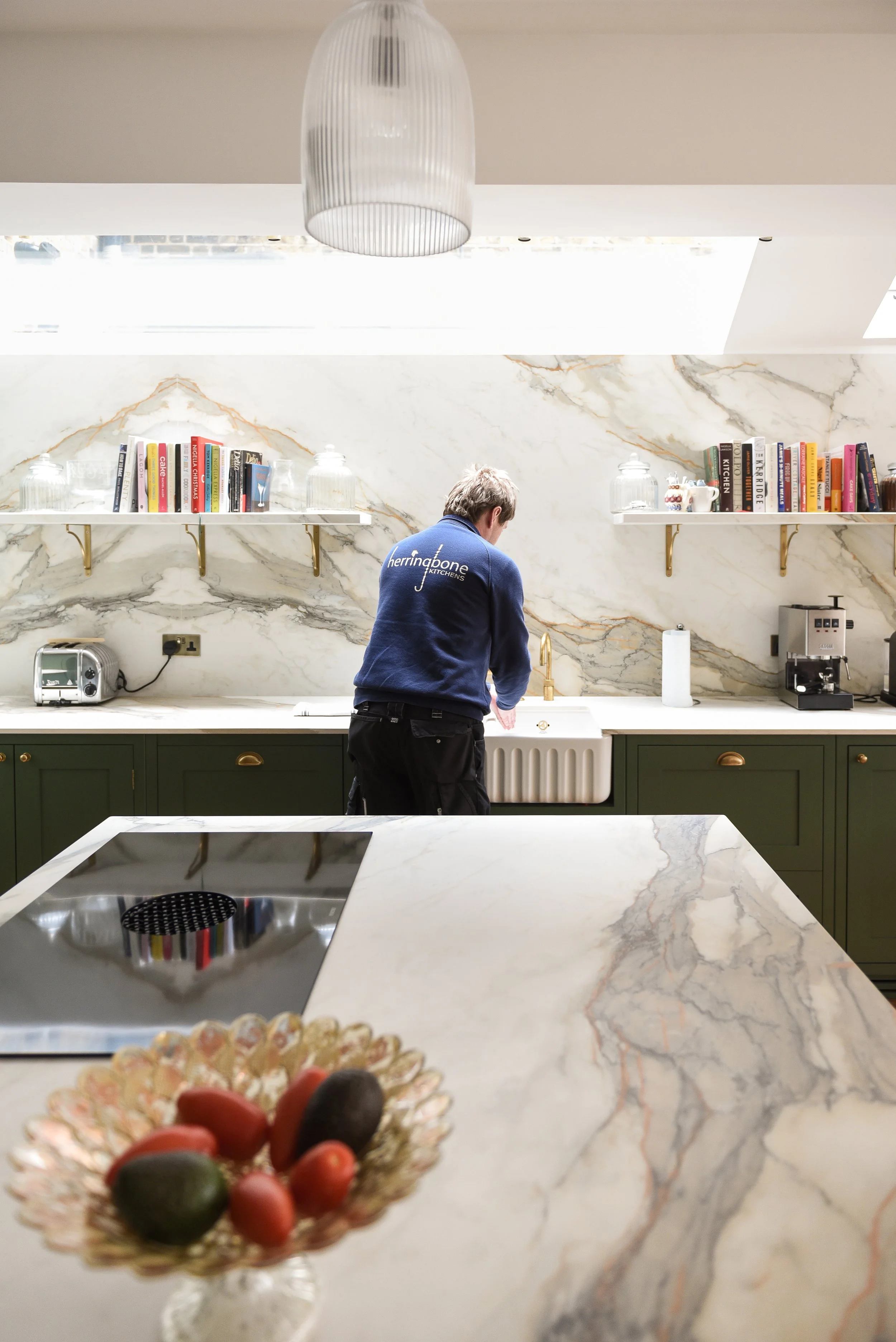 A person wearing a blue Herringbone Kitchens jacket is seen in a modern kitchen with marble countertops and a marble backsplash, seen from behind, standing at a sink.