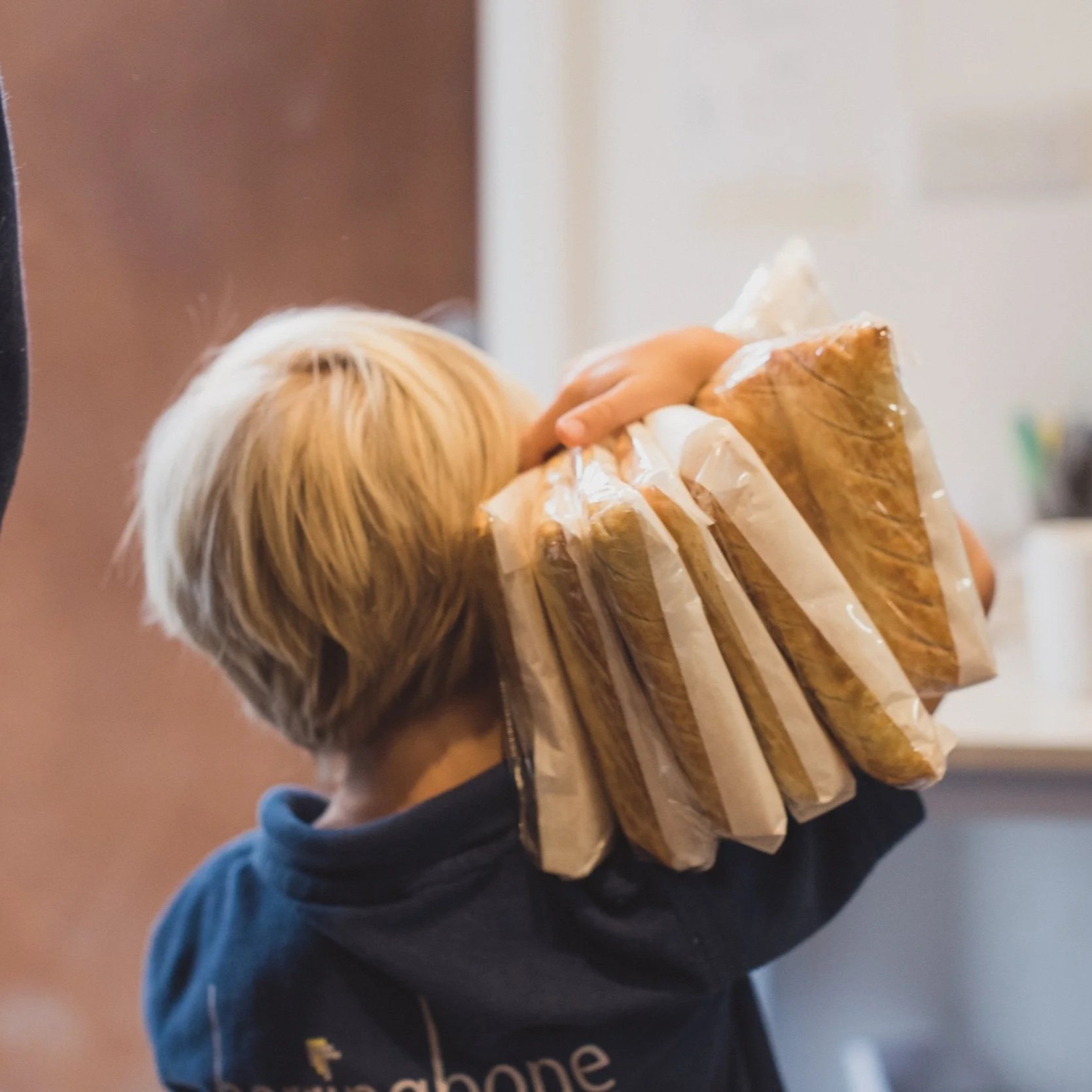 Person with blond hair wearing a dark hoodie carries a paper bag filled with a sandwich and a smoothie.
